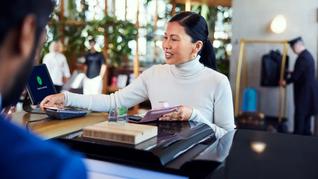 Una mujer en el mostrador de recepción de un hotel pagando su estancia tocando una tarjeta en el terminal de Adyen.