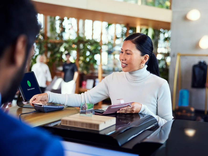 Una mujer en el mostrador de recepción de un hotel pagando su estancia tocando una tarjeta en el terminal de Adyen.