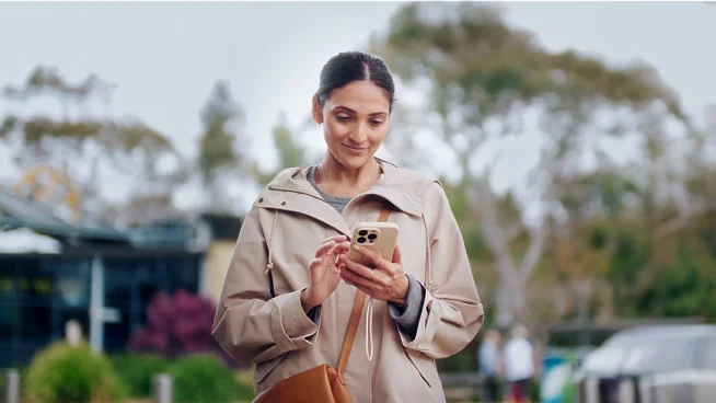 Smiling woman in a beige coat using a smartphone outside.