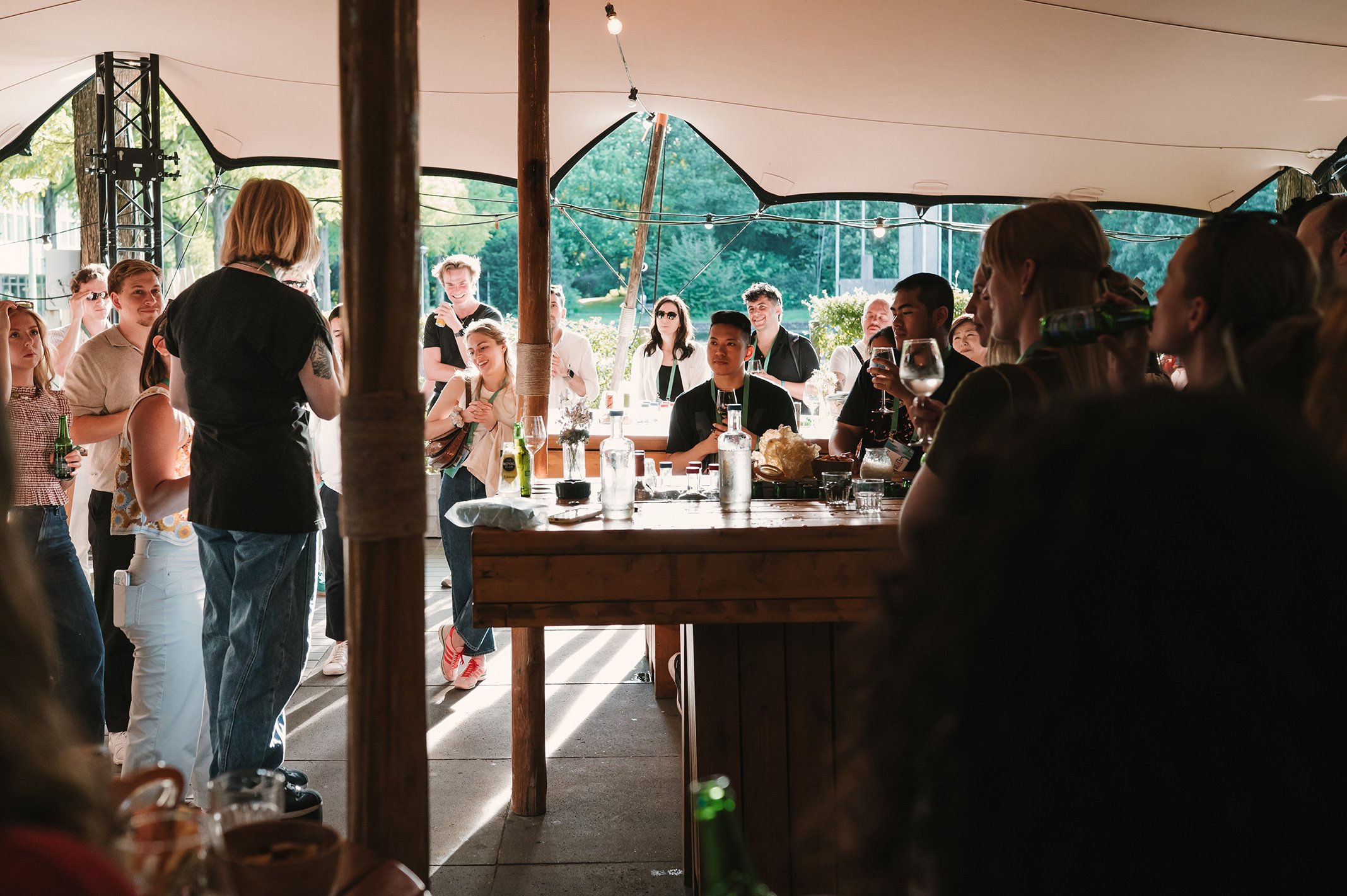 Guests gather at an outdoor event with beverages on a sunny day, under a canopy.