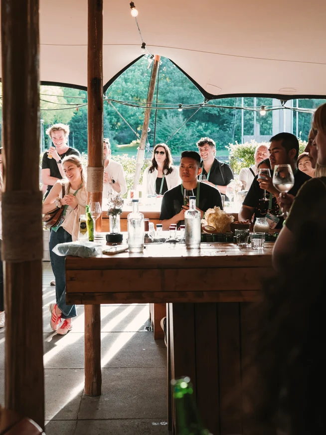 Guests gather at an outdoor event with beverages on a sunny day, under a canopy.