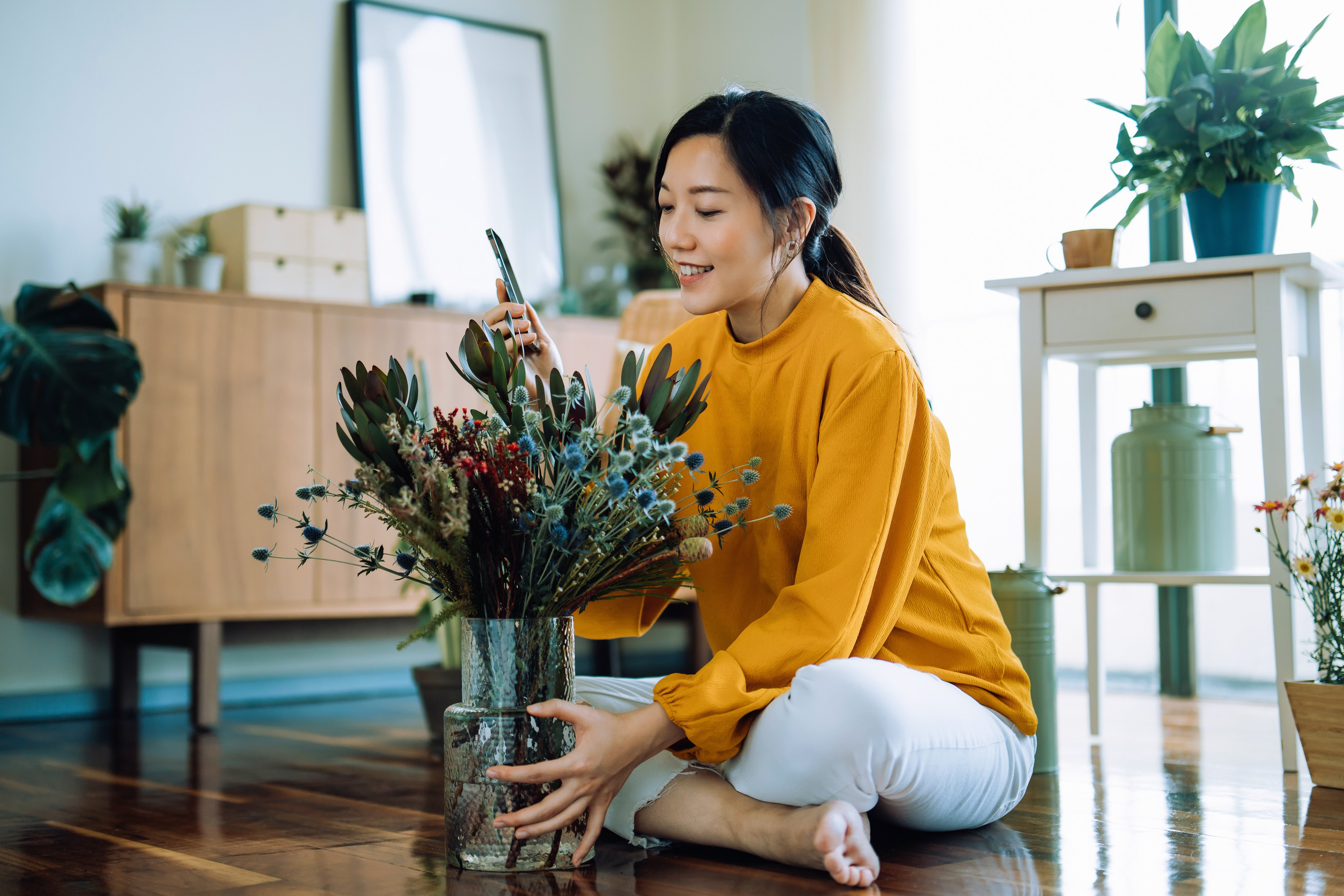 A woman arranging flowers indoors with natural light, in a space decorated with plants and furniture.