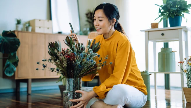 A woman arranging flowers indoors with natural light, in a space decorated with plants and furniture.
