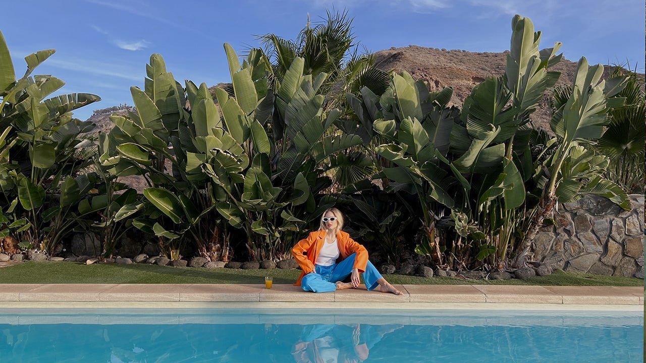 Kvinde i orange jakke og blå bukser sidder ved en pool med tropiske planter i baggrunden.