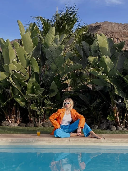 Kvinde i orange jakke og blå bukser sidder ved en pool med tropiske planter i baggrunden.