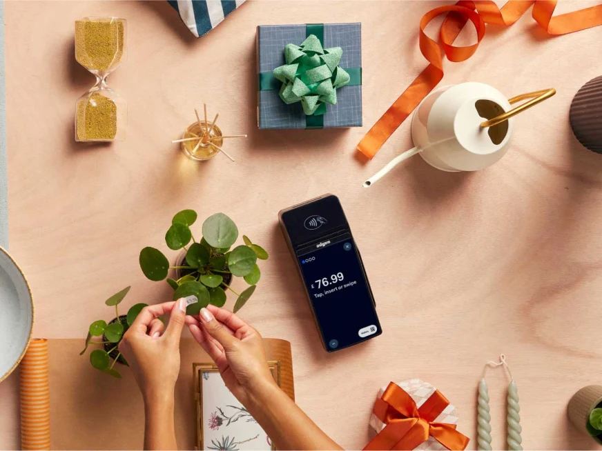 Hands wrapping a plant stem with a bow, next to a smartphone showing Adyen payment of $76.99.