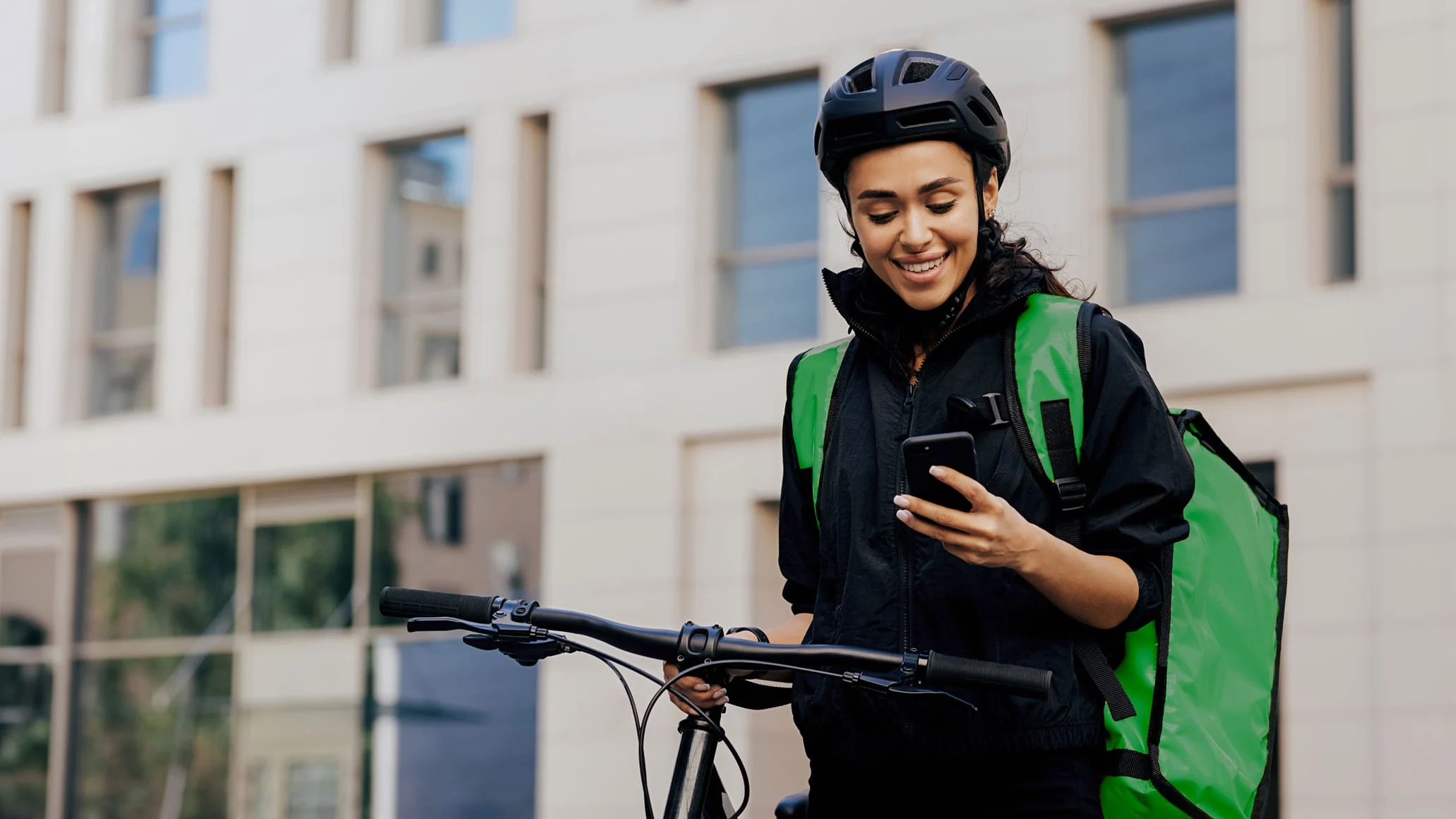 A woman cyclist with a helmet and backpack using her phone beside a bicycle outdoors.