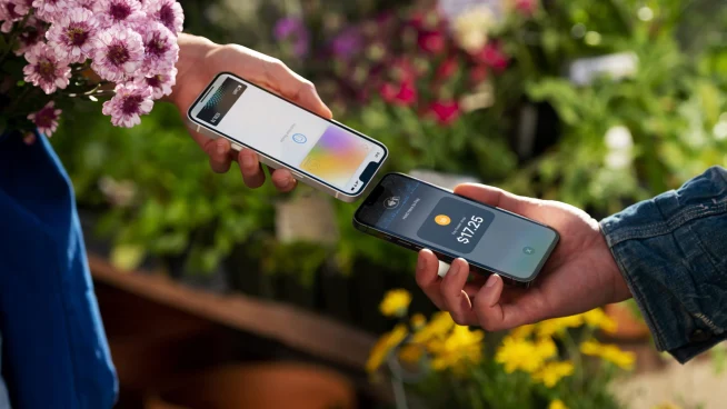 Two hands holding smartphones, one performing a contactless payment to the other amidst plants.