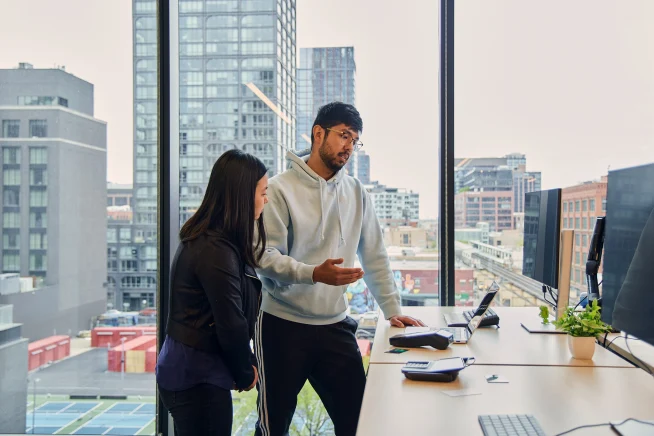 two collegues standing behind their desk, having a conversation while looking at their laptop 