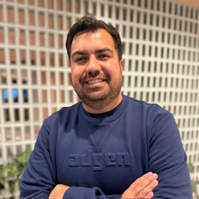 Adyen employee in a navy sweater smiling with crossed arms in front of a modern office wall.