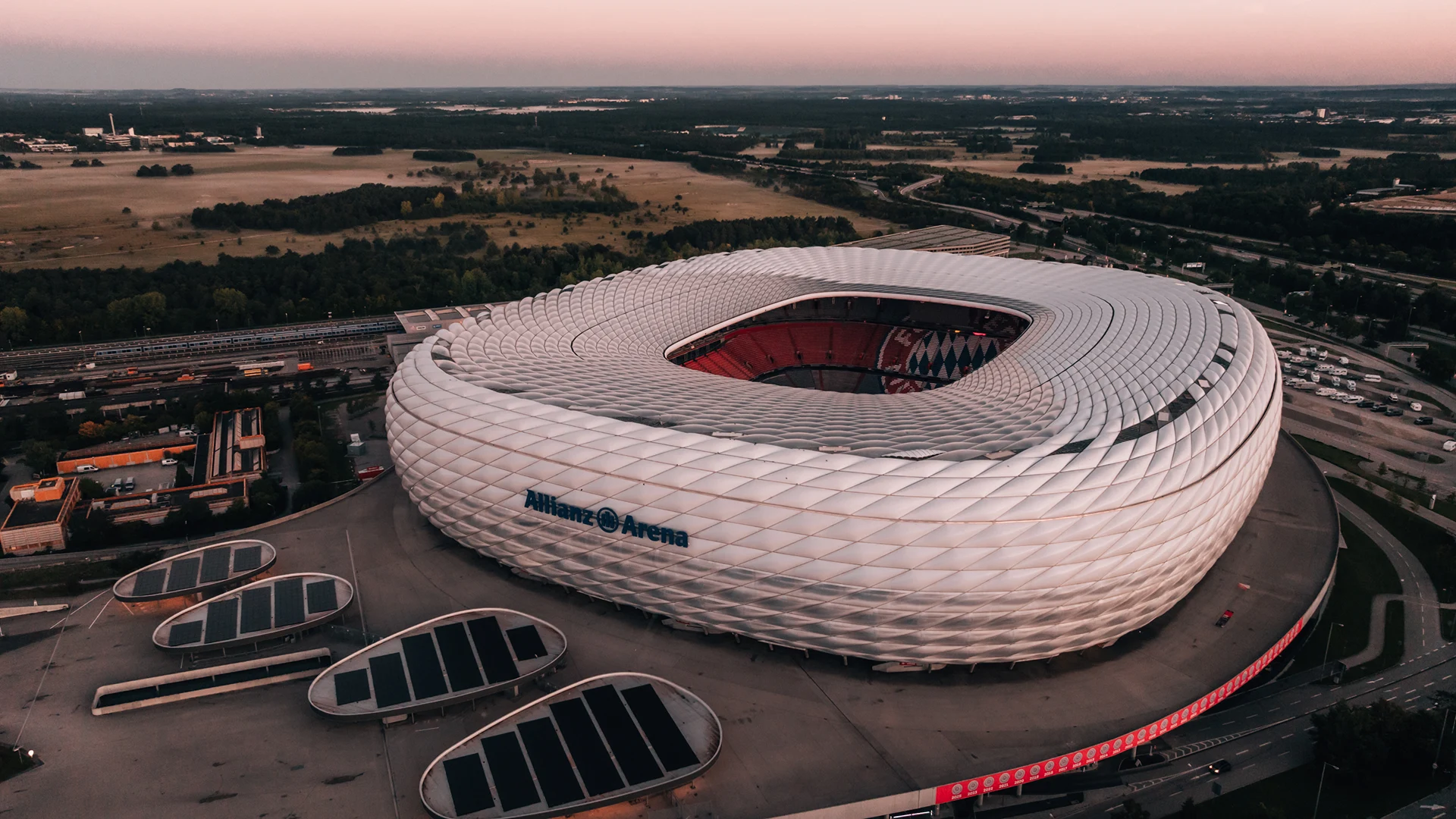 A large, modern stadium with a white exterior, seen from above, during dusk.
