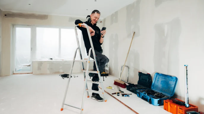 Ein Tooltime-Mitarbeiter auf einer Leiter in einem leeren Raum blickt auf sein Smartphone.