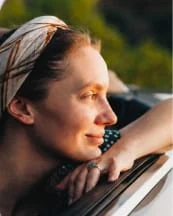 A woman leaning on a car window, enjoying the outdoors, with sunlight highlighting her face.