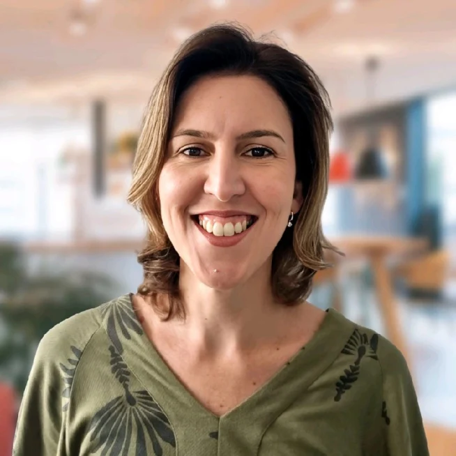 A woman smiling in an indoor setting, possibly an office or cafe, with a blurred background.