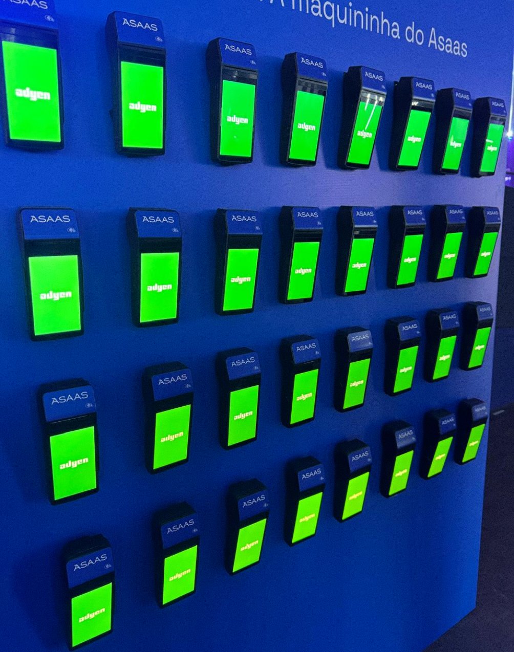 Várias maquininhas de cartão Adyen em uma parede azul, com telas verdes exibindo o status da transação.
