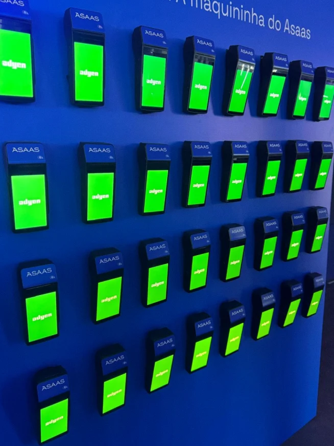 Várias maquininhas de cartão Adyen em uma parede azul, com telas verdes exibindo o status da transação.