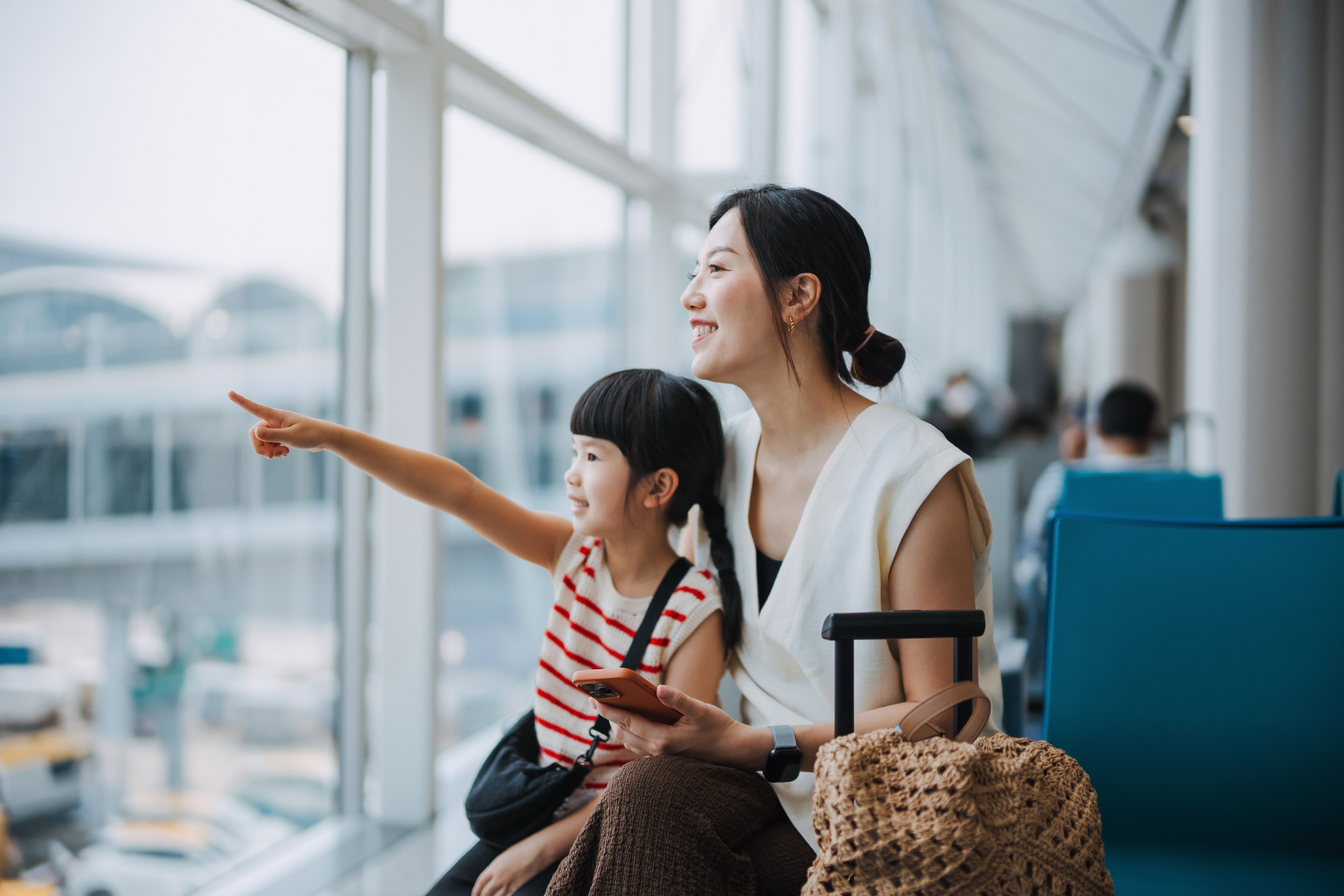 A woman and girl at an airport terminal, with the girl pointing, while a baggage trolley with a woven bag is visible.