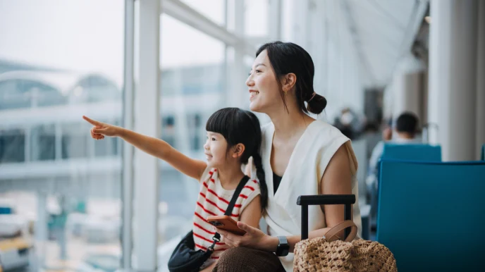 Woman and girl at an airport terminal, the girl pointing, with luggage and a woven bag on a trolley nearby.