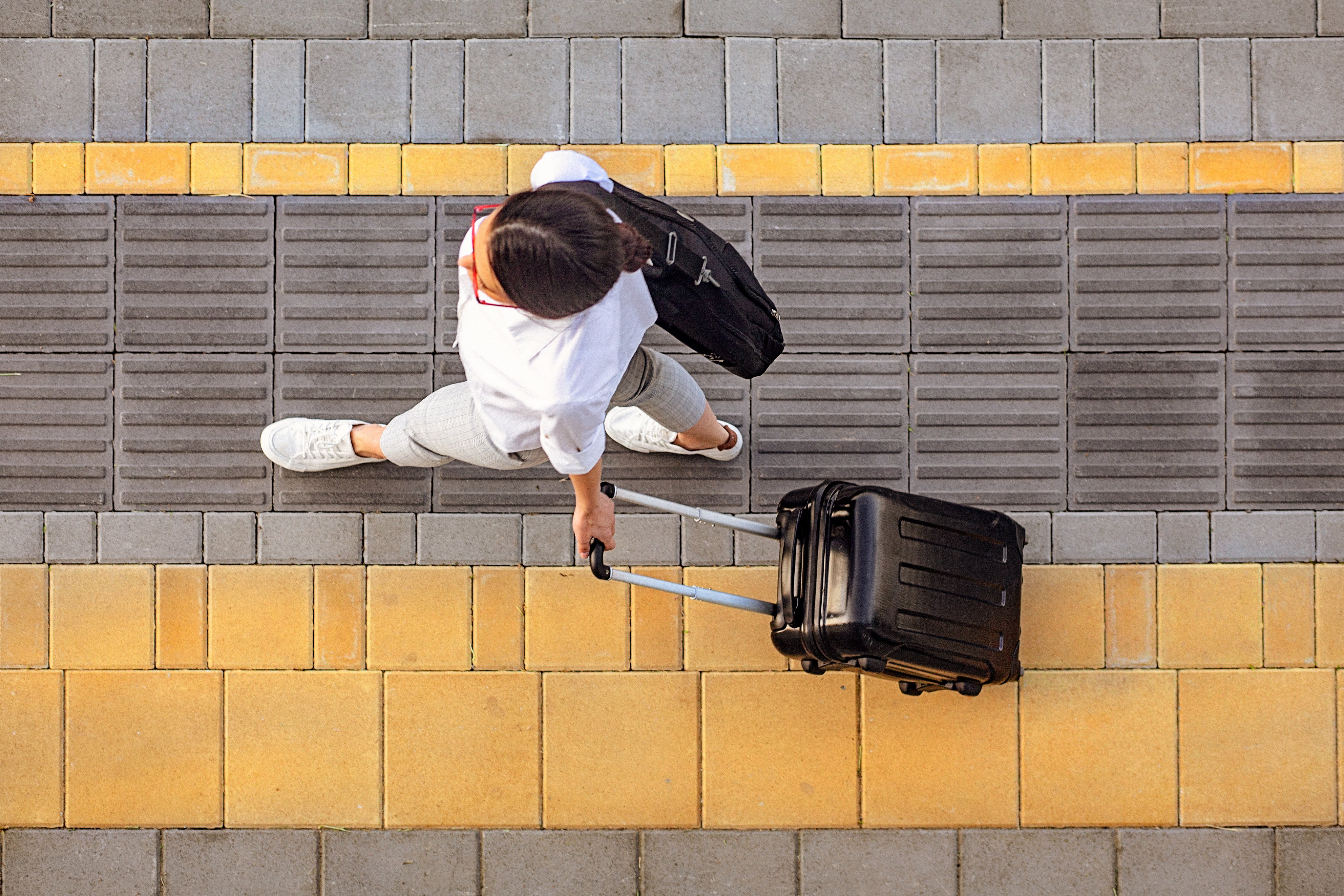 A traveler with a suitcase walking on a sidewalk with yellow and grey tiles, carrying a black backpack.