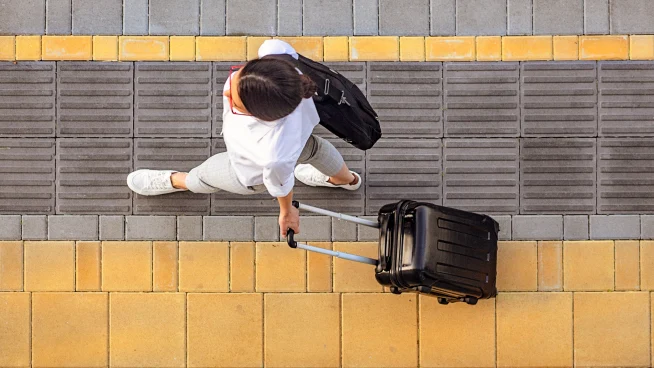 A traveler with a suitcase walking on a sidewalk with yellow and grey tiles, carrying a black backpack.