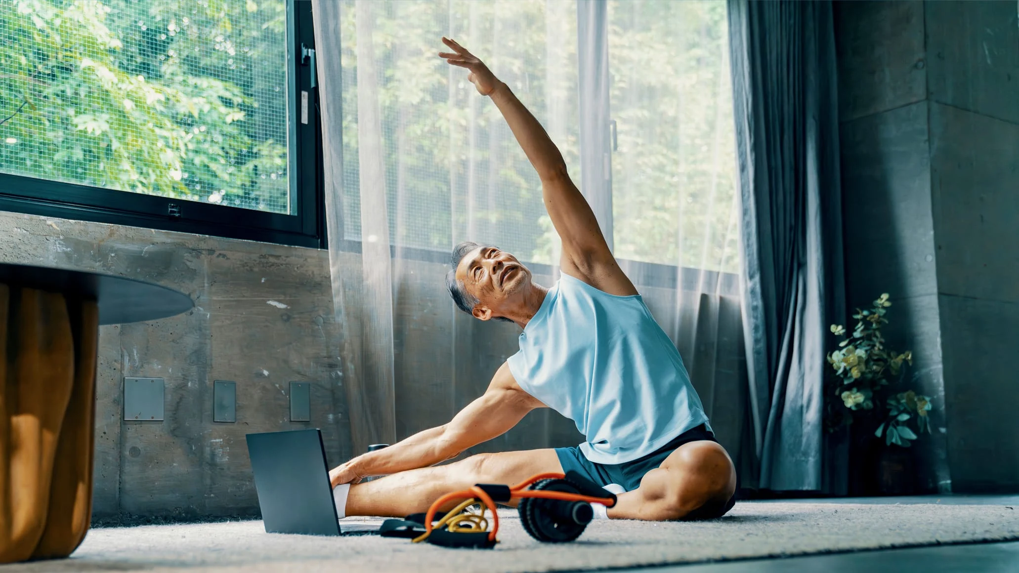 A person stretches on the floor near a laptop in a bright indoor space with large windows and curtains.
