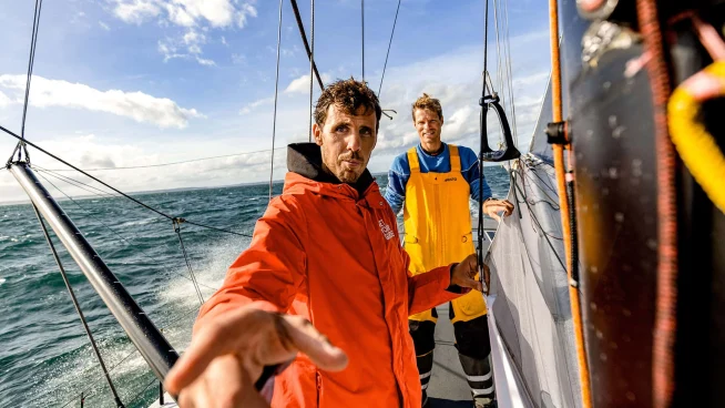 Two men on a sailing boat with one extending his hand towards the camera.