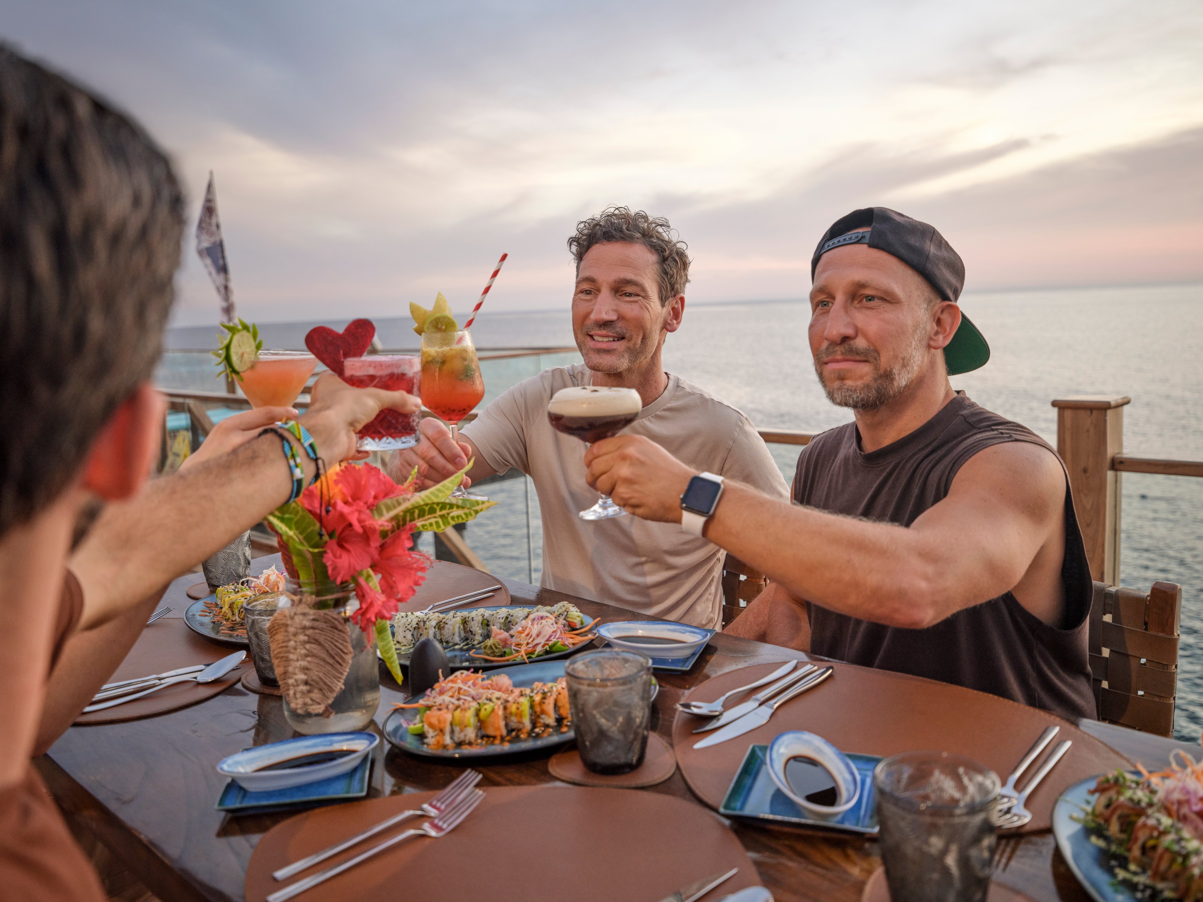 Menschen beim gemeinschaftlichen Essen am Strand, mit bunten Cocktails und Blick aufs Meer