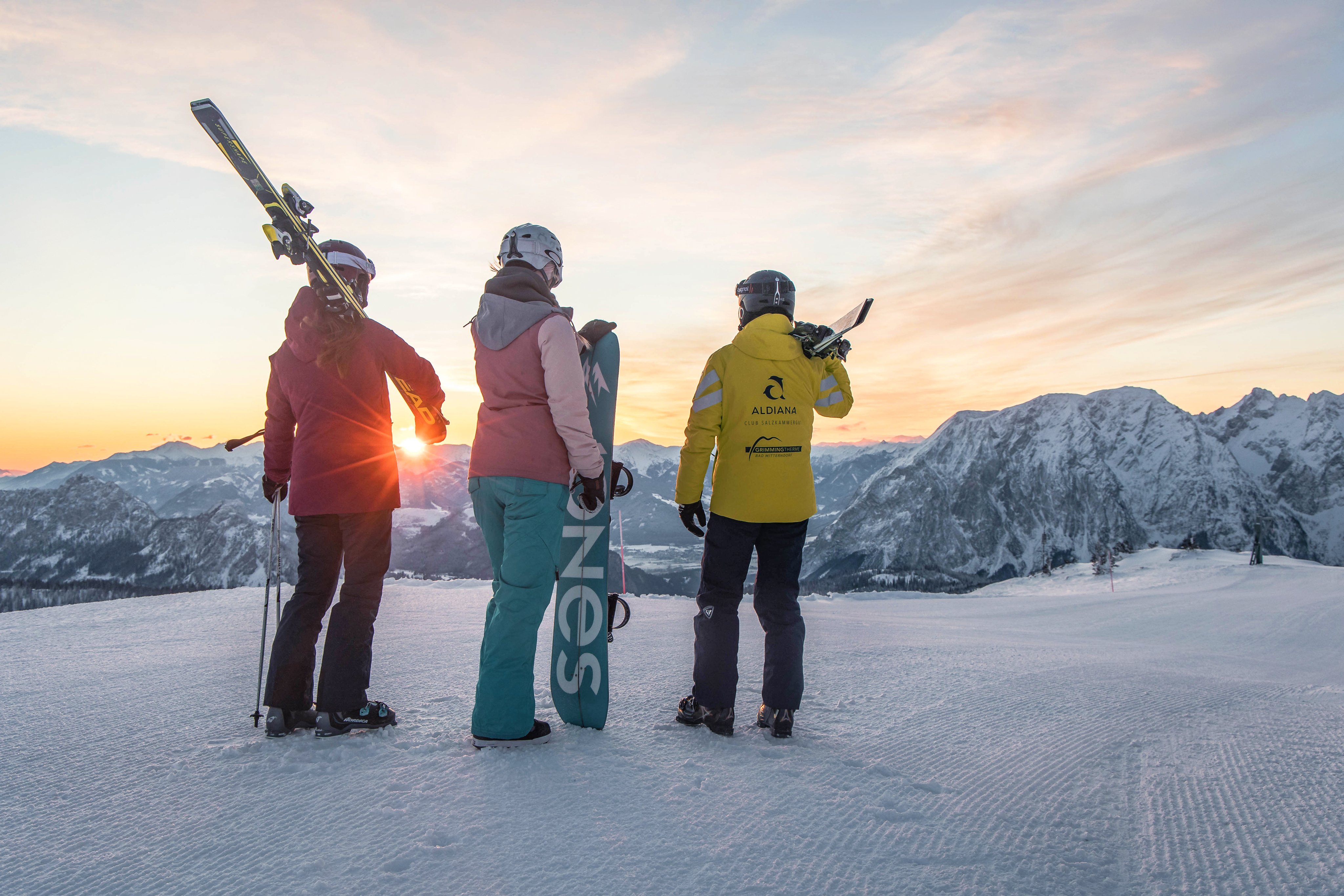 Drei Skifahrer auf verschneitem Berggipfel bei Sonnenuntergang, Blick auf die Alpen.