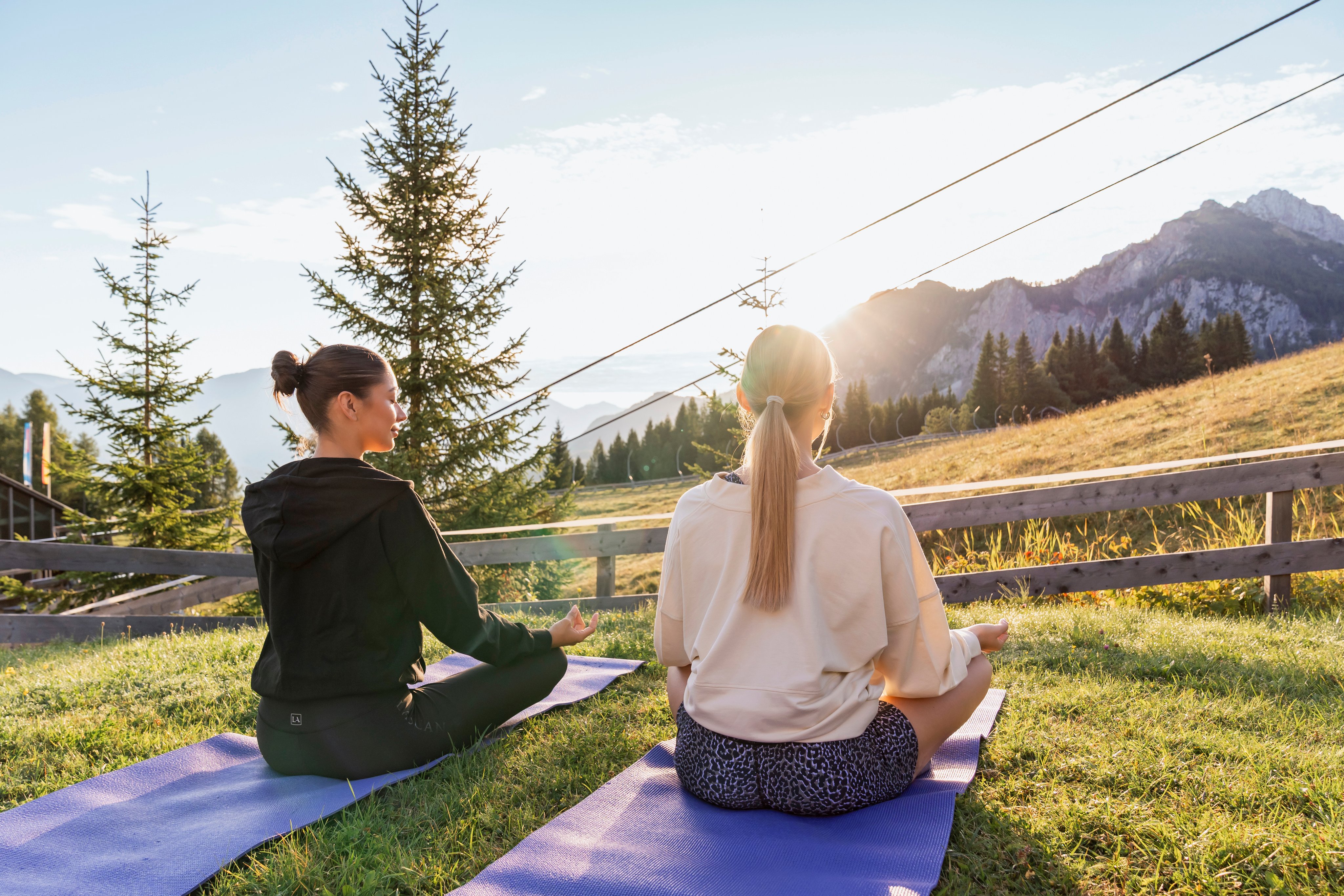 Zwei Frauen sitzen auf Yogamatten im Grünen und genießen den Sonnenuntergang in den Bergen