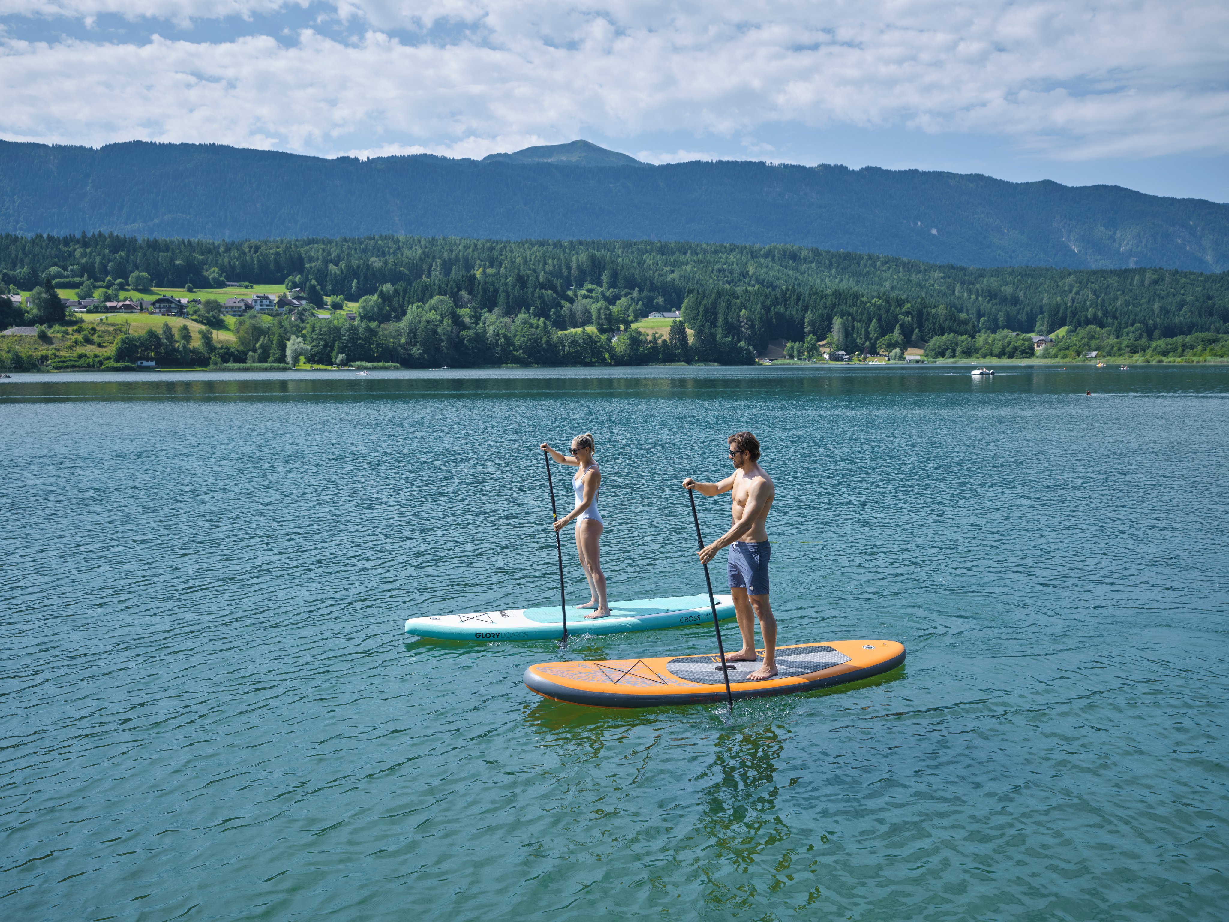 Zwei Kinder auf Stand-Up-Paddle-Boards auf einem ruhigen See mit Bergen im Hintergrund