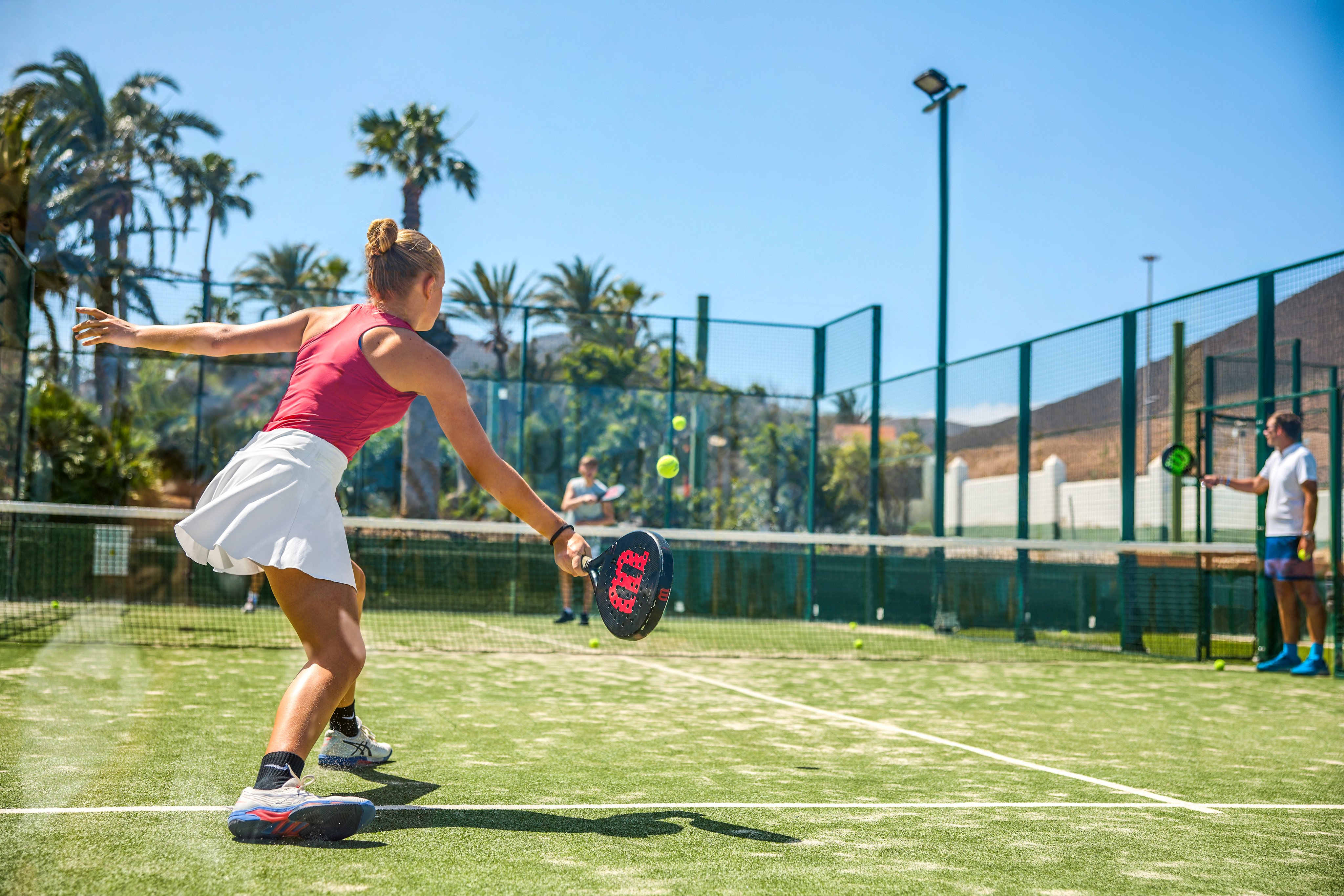 Frau beim Tennis auf einem grünen Platz, Palmen im Hintergrund, sonniges Wetter