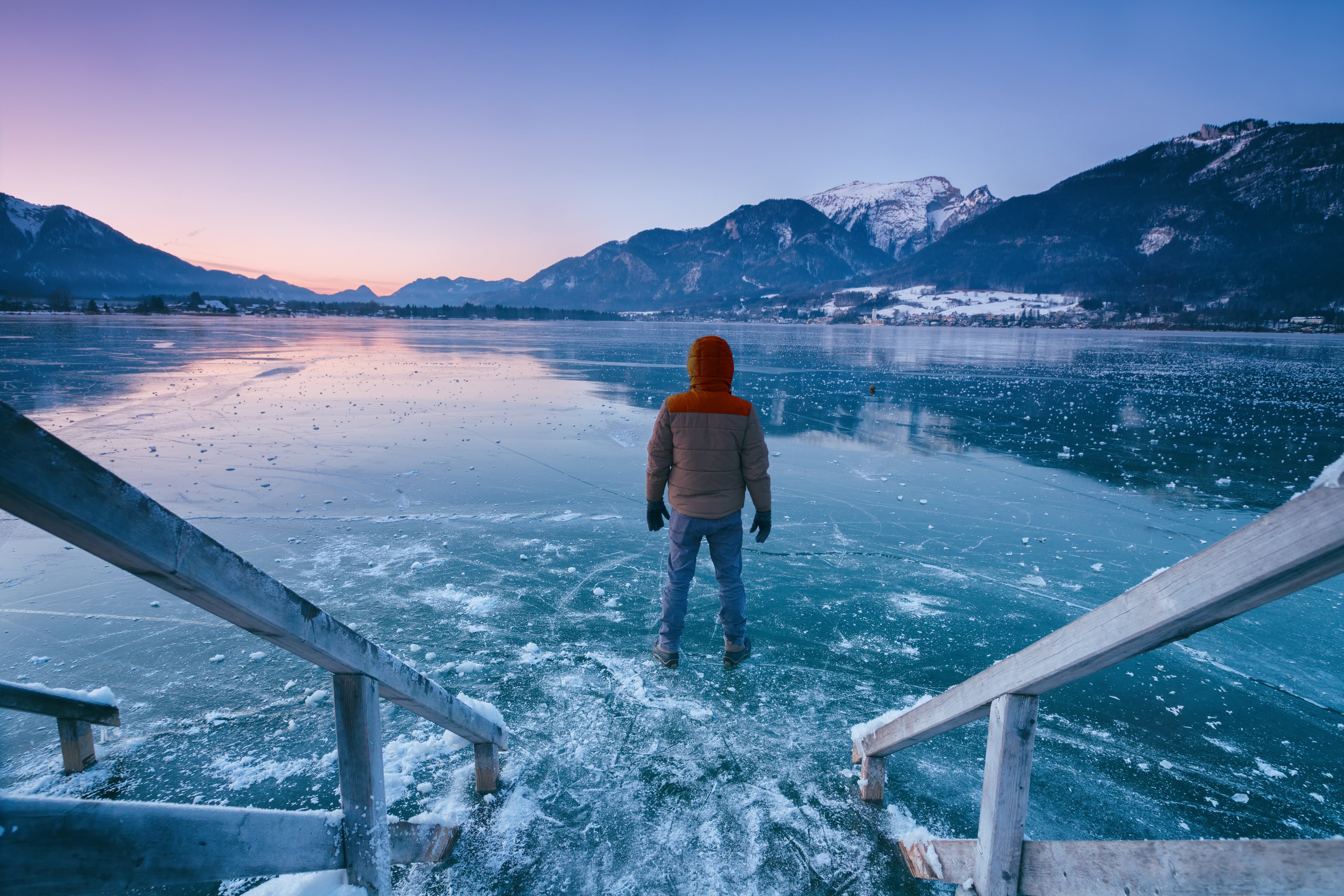 Person steht auf einem Eissee vor Bergen, Wasser spiegelt den Himmel, Sonnenuntergang im Hintergrund