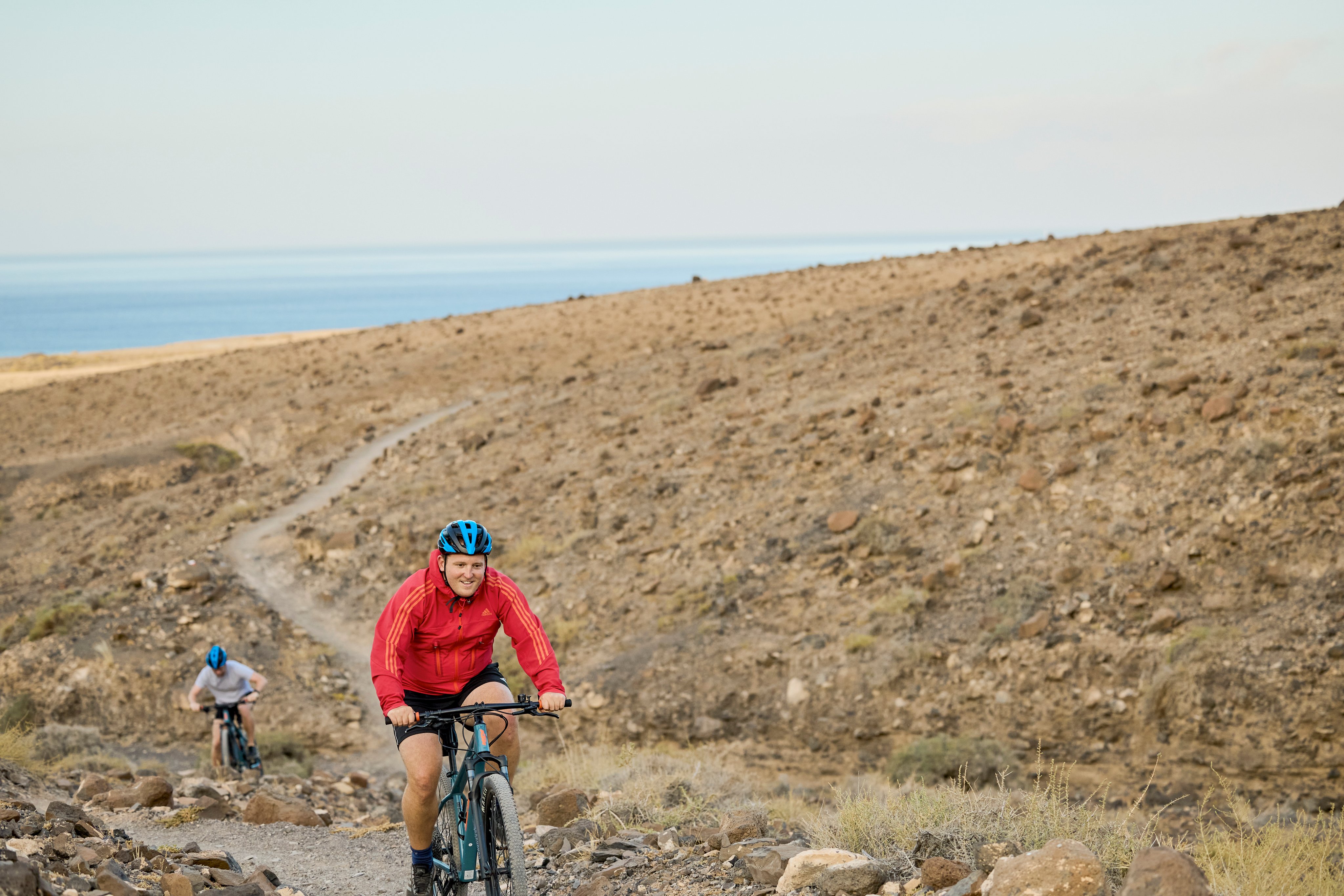 Biker auf einem felsigen Wanderpfad in einer kargen, bergigen Landschaft mit Blick aufs Meer
