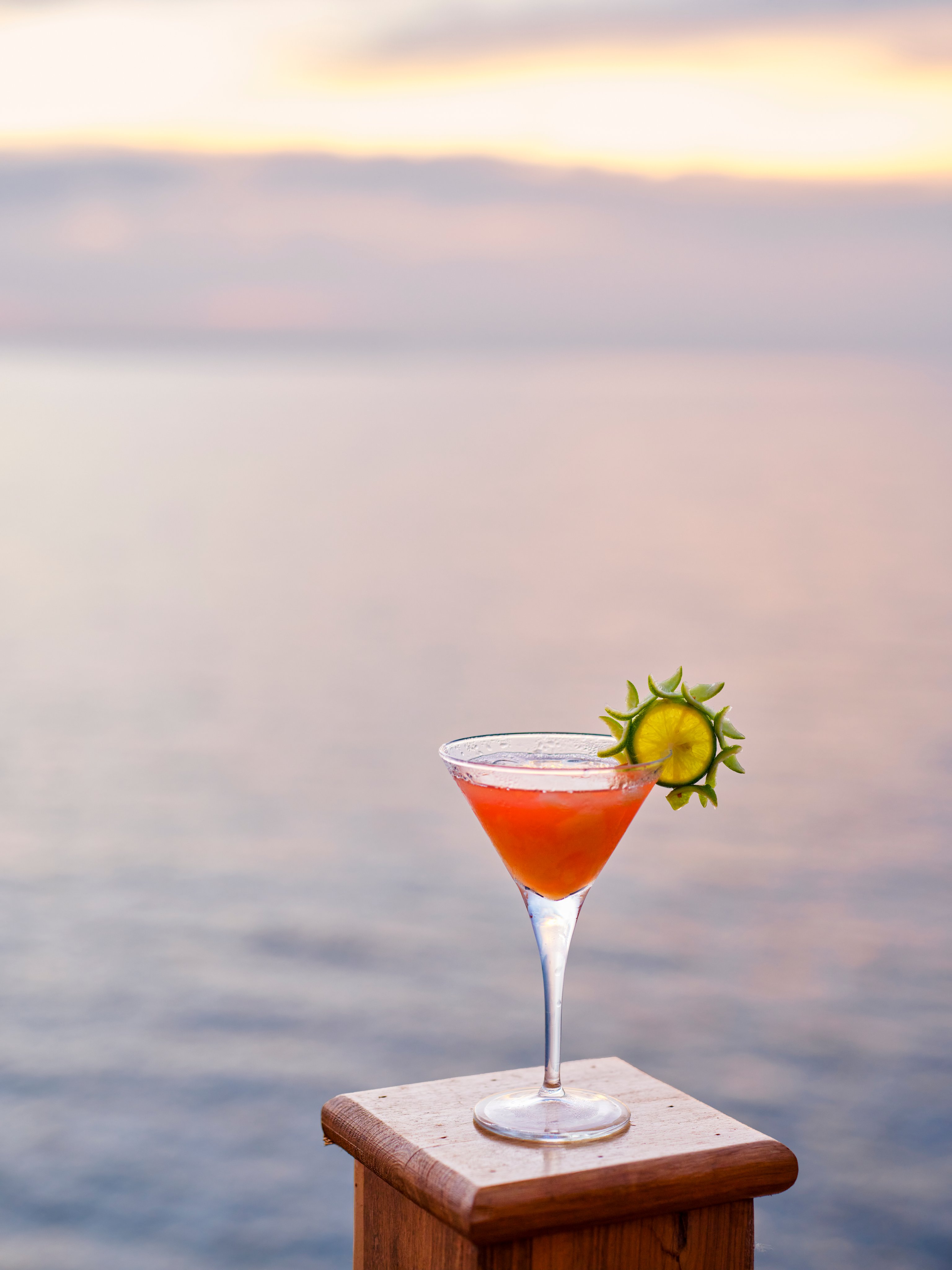 Ein gekühlter Fruchtcocktail mit Gurkenscheibe auf einem Holzuntergrund am Meer bei Sonnenuntergang
