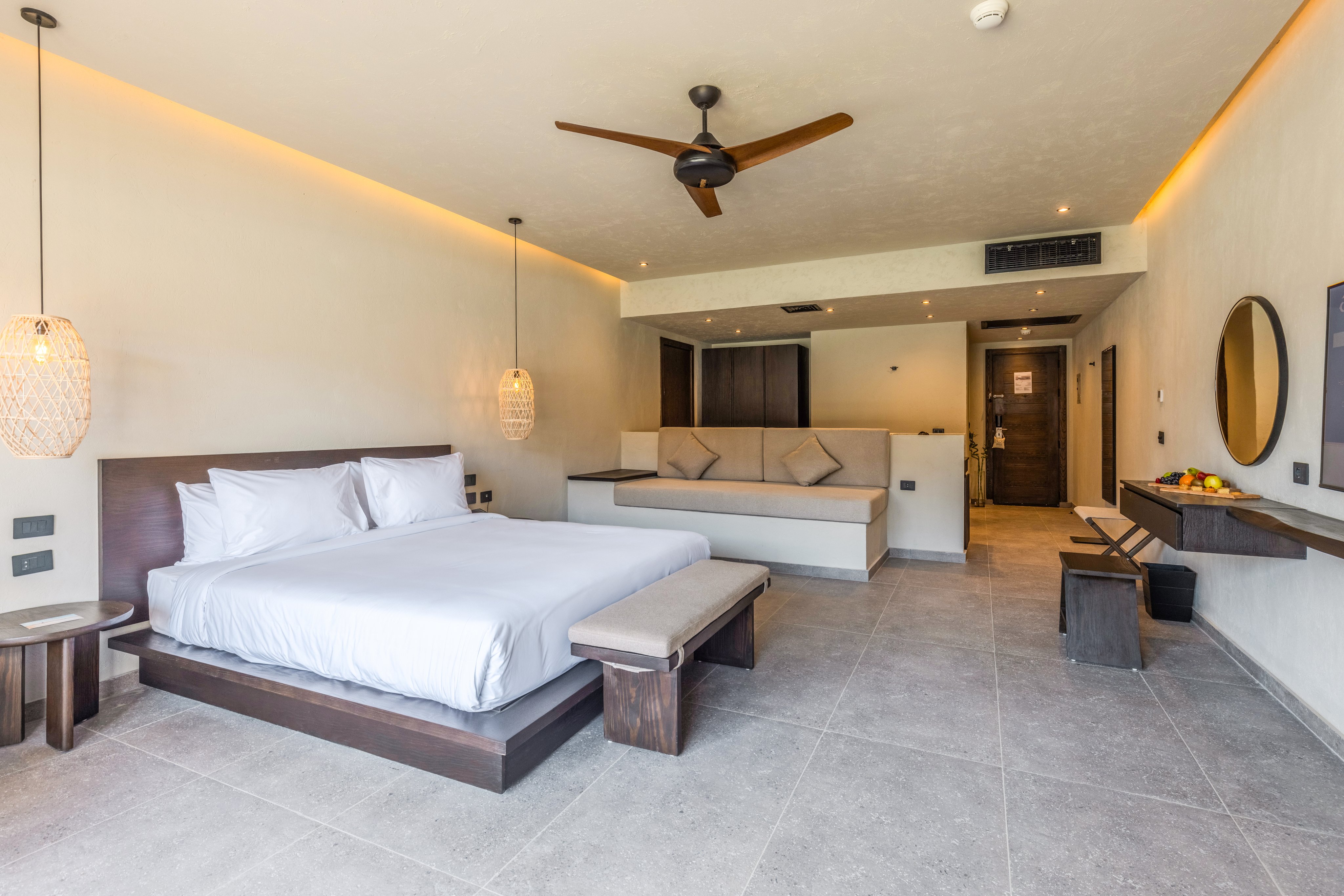 Modernes Hotelzimmer mit Bett, Sofa, Beleuchtung, Deckenventilator und TV in deutscher Sprache.