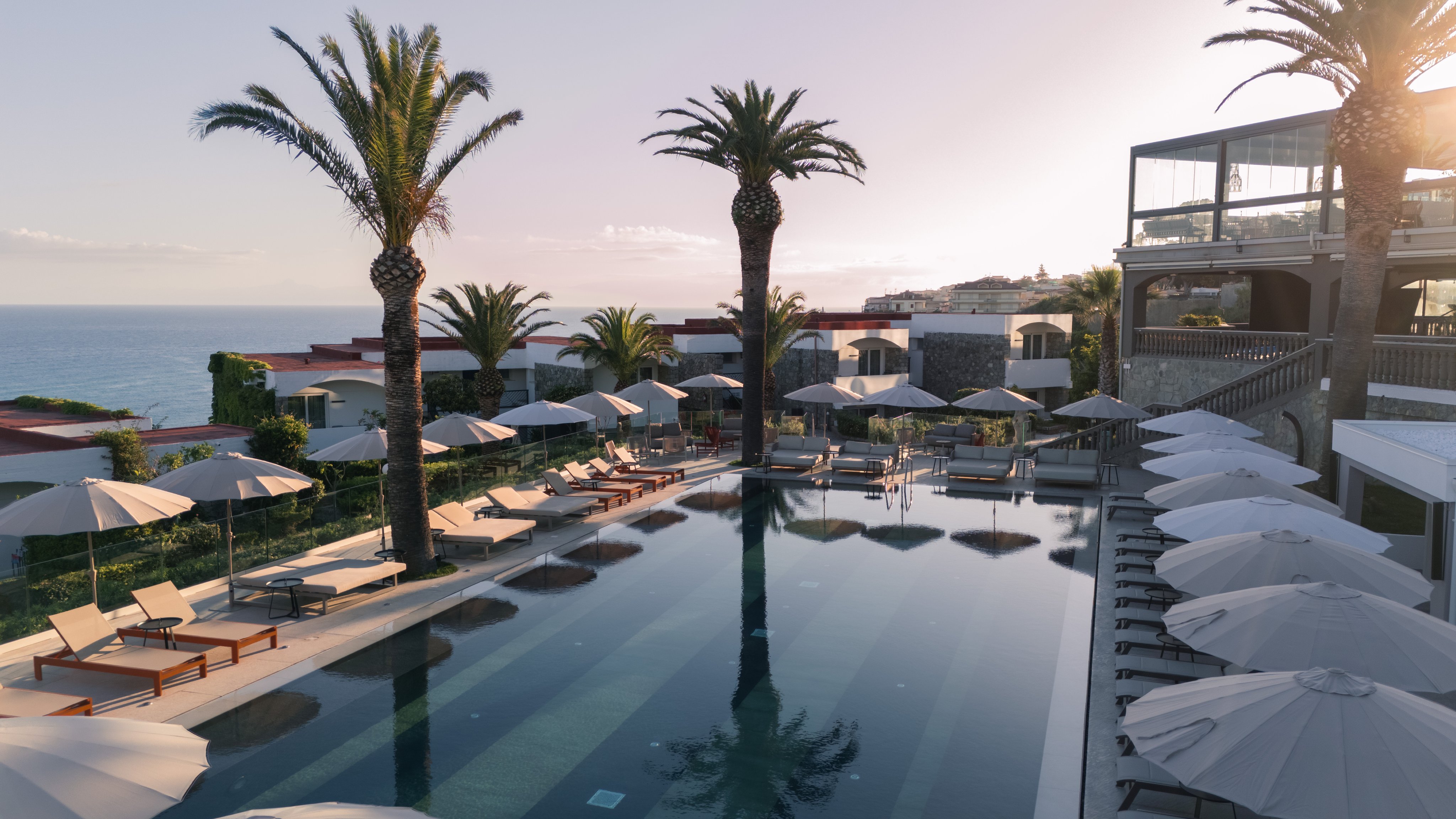 Luxuriößer Poolbereich mit Palmen und Liegen, Blick auf Meer bei Sonnenuntergang