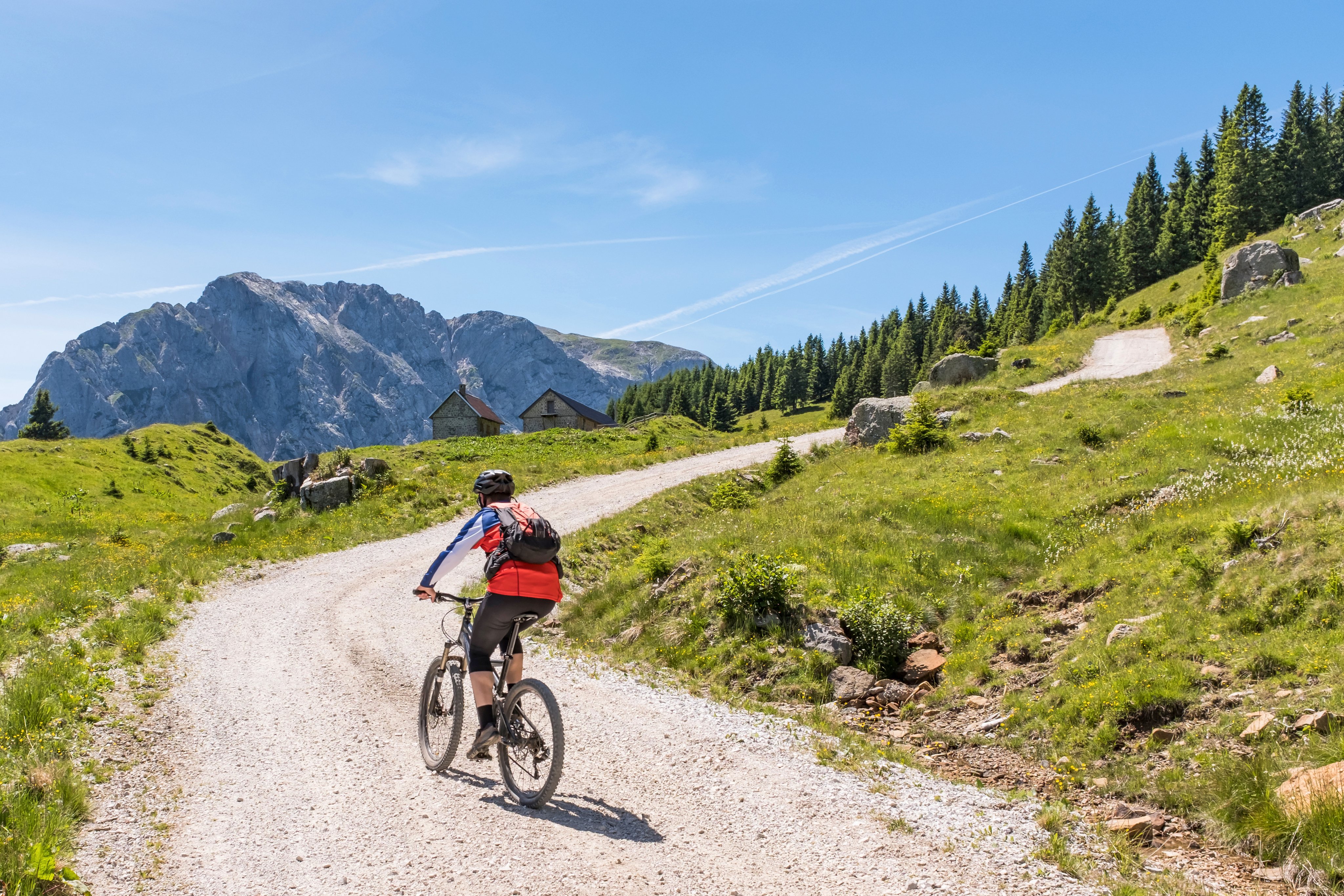 Eine Person fährt mit dem Mountainbike auf einer gewundenen Schotterstraße in den Bergen, umgeben von grüner Landschaft und Bergen im Hintergrund.