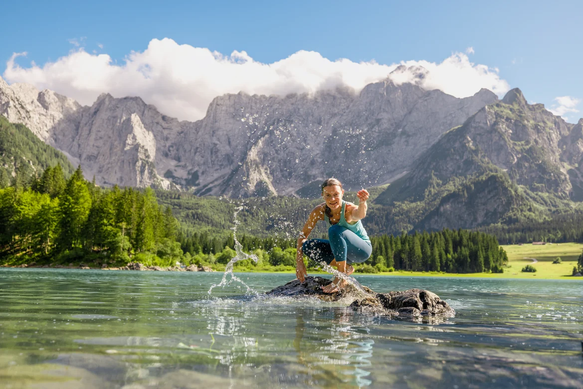 Person beim Balancieren auf einem Stein im klaren Bergsee vor einer beeindruckenden Bergkulisse
