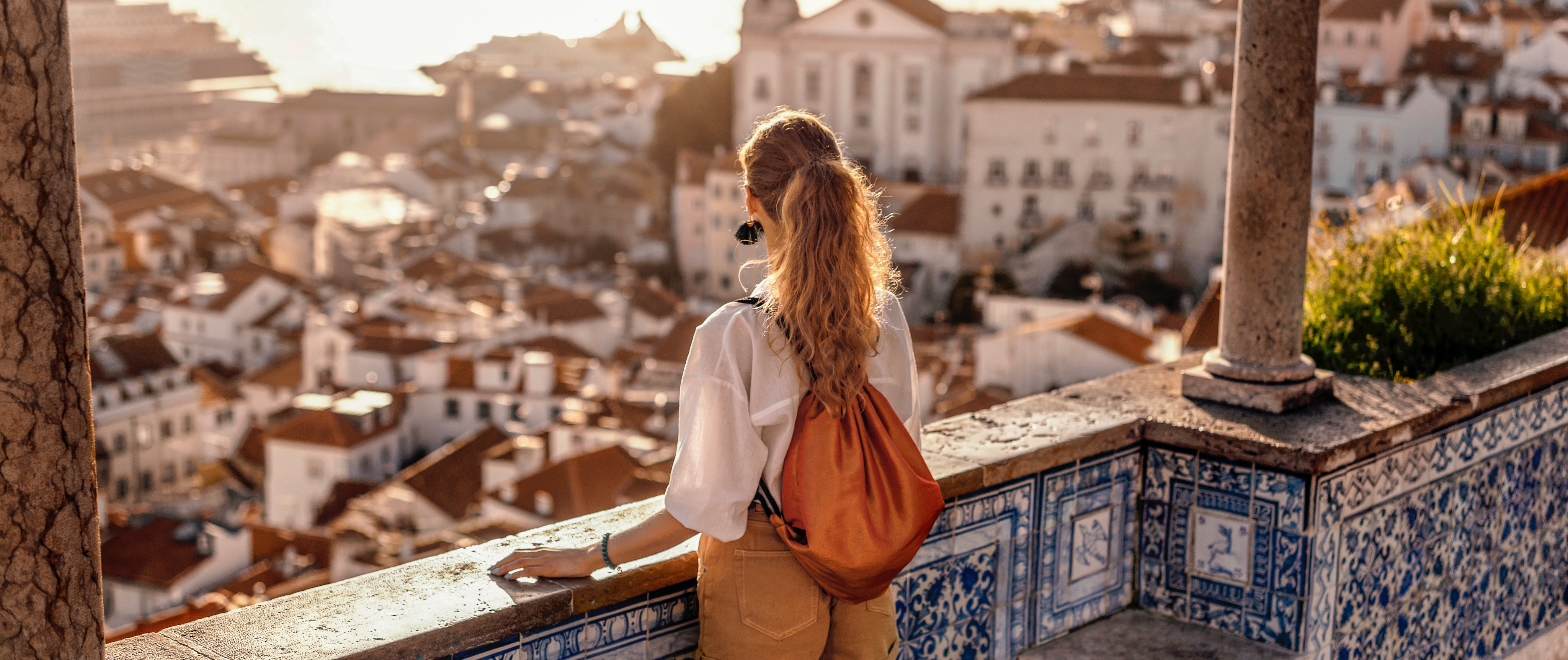 Eine Frau mit Locken schaut auf eine mediterrane Stadt von einer Terrasse aus, im Sonnenlicht