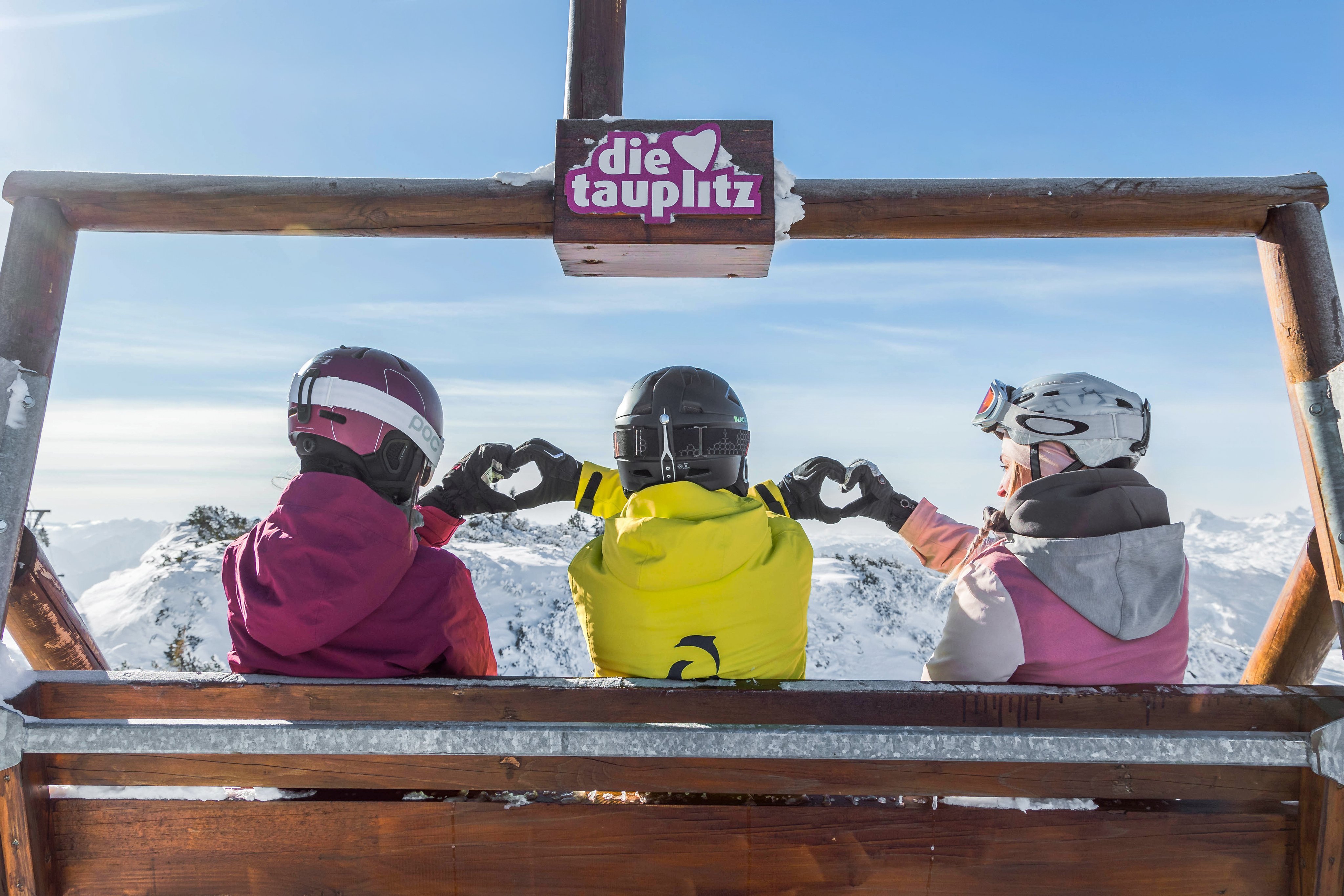 Drei Kinder in Skiausrüstung auf einer Bergbank mit Blick auf schneebedeckte Berge