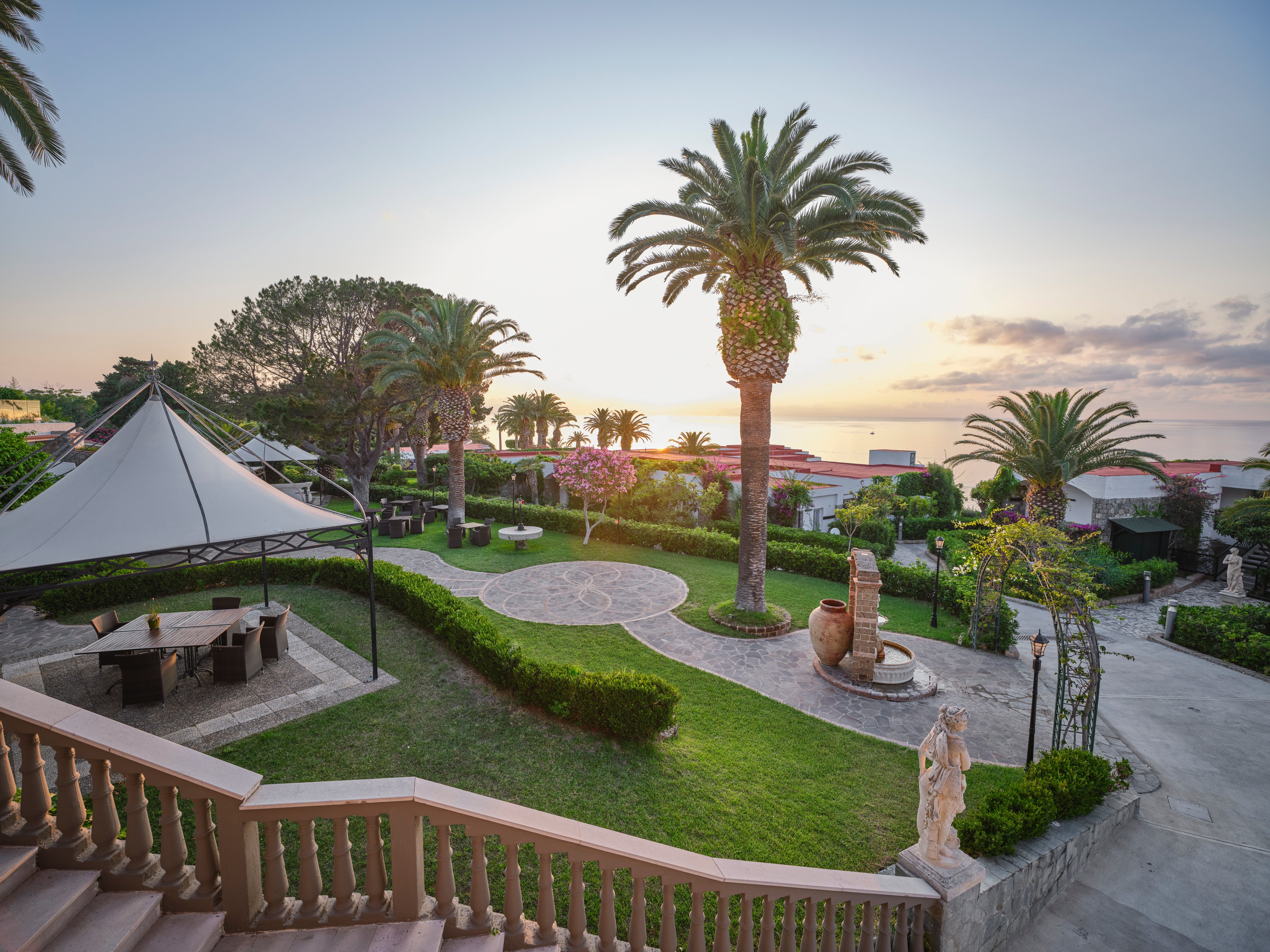 Blick auf einen gepflegten, grünen Hotelgarten mit Palmen und Sitzgelegenheiten bei Sonnenuntergang