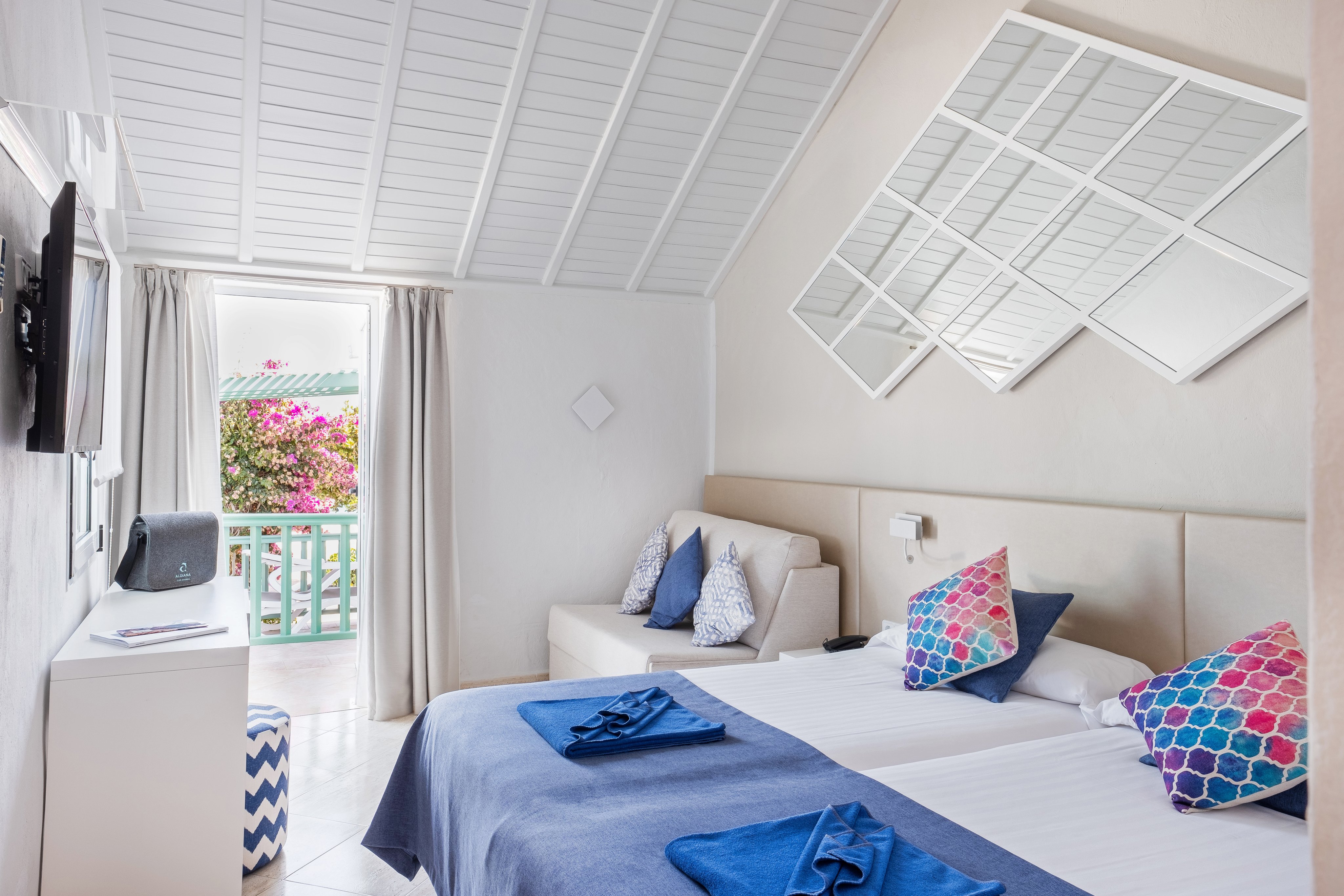 Ein helles Hotelzimmer mit Doppelbett, zwei Nachtschränkchen, Couch, TV, Balkon und große Fenster