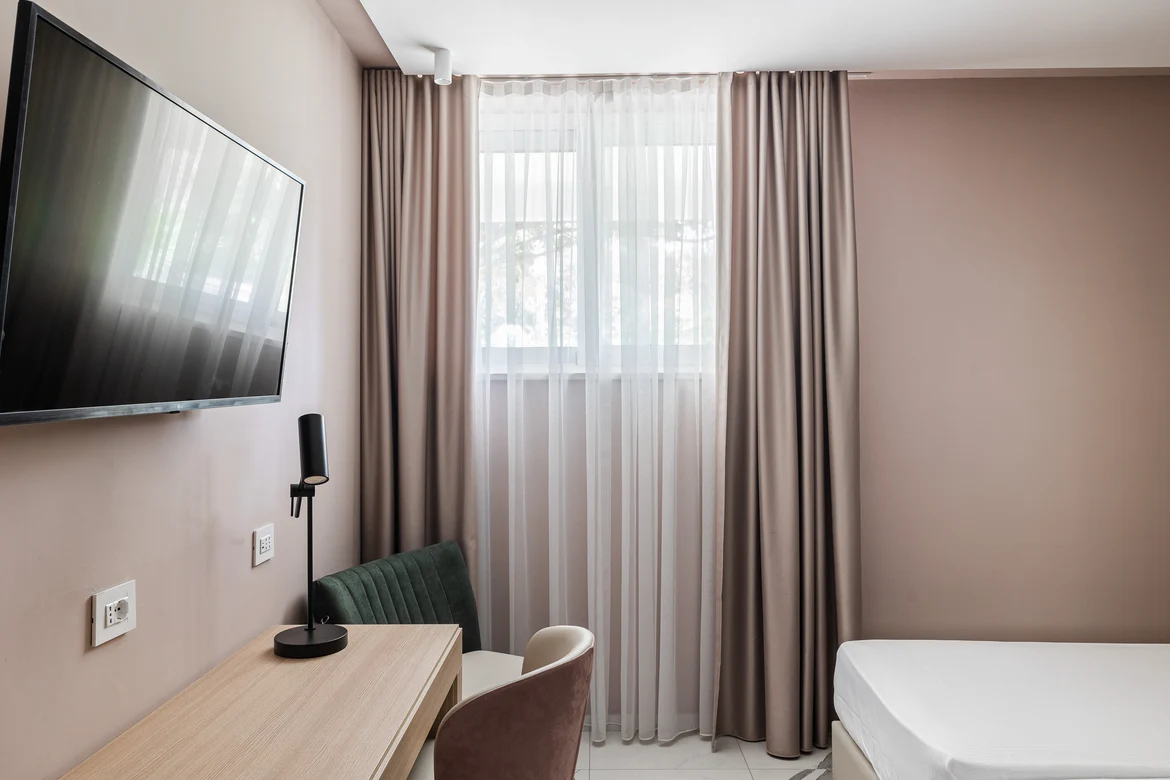 Elegantes Hotelzimmer mit Bett, Schreibtisch, Stuhl, Fernseher und Fenster mit Vorhängen