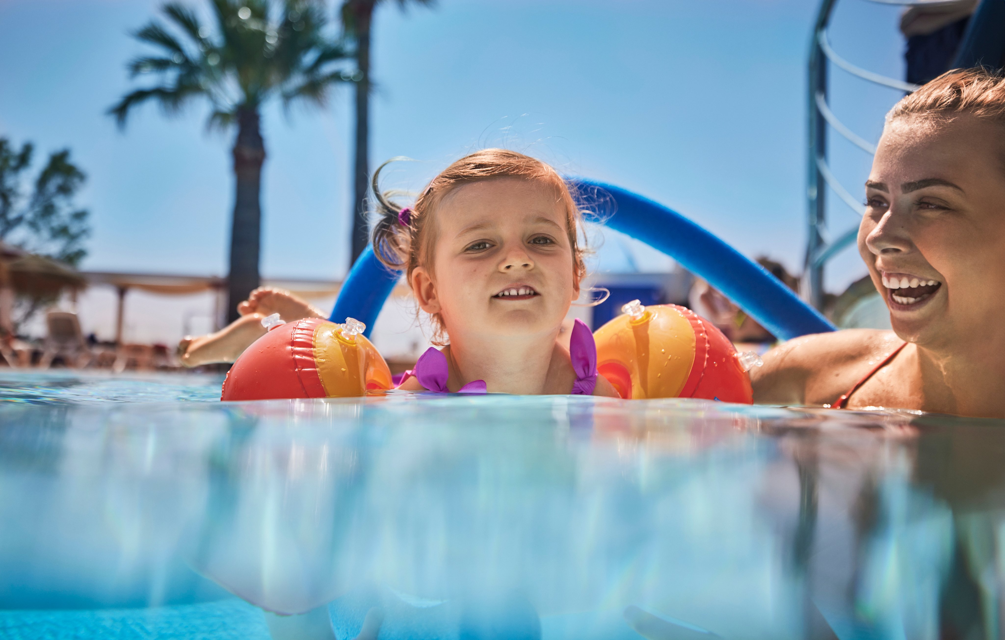Kinder beim Schwimmen im Wasser mit Schwimmhilfen und Palmen im Hintergrund