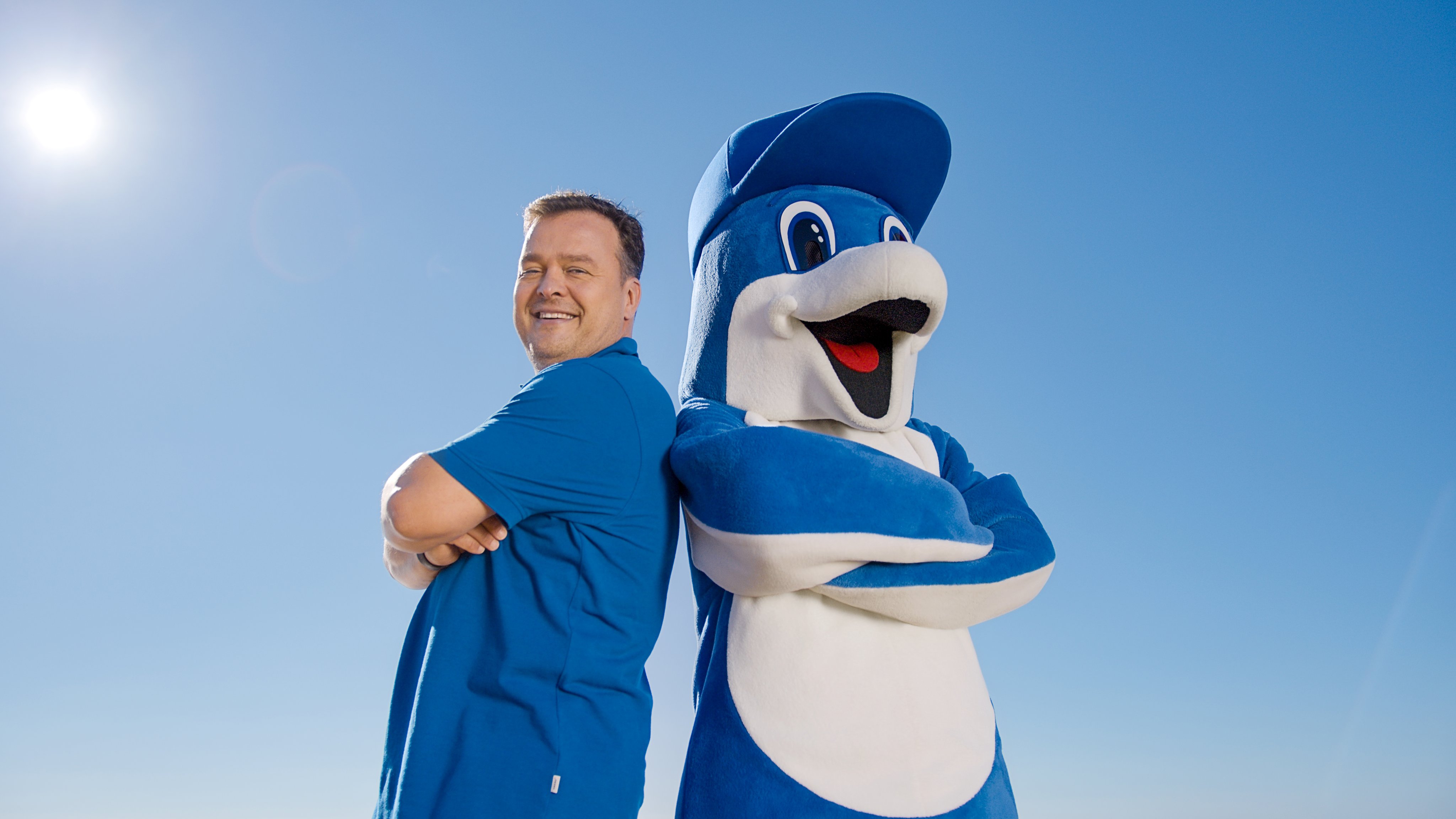 Ein Mann steht mit verschränkten Armen neben einer mascottengestalt von Aldiana im blauen Outfit