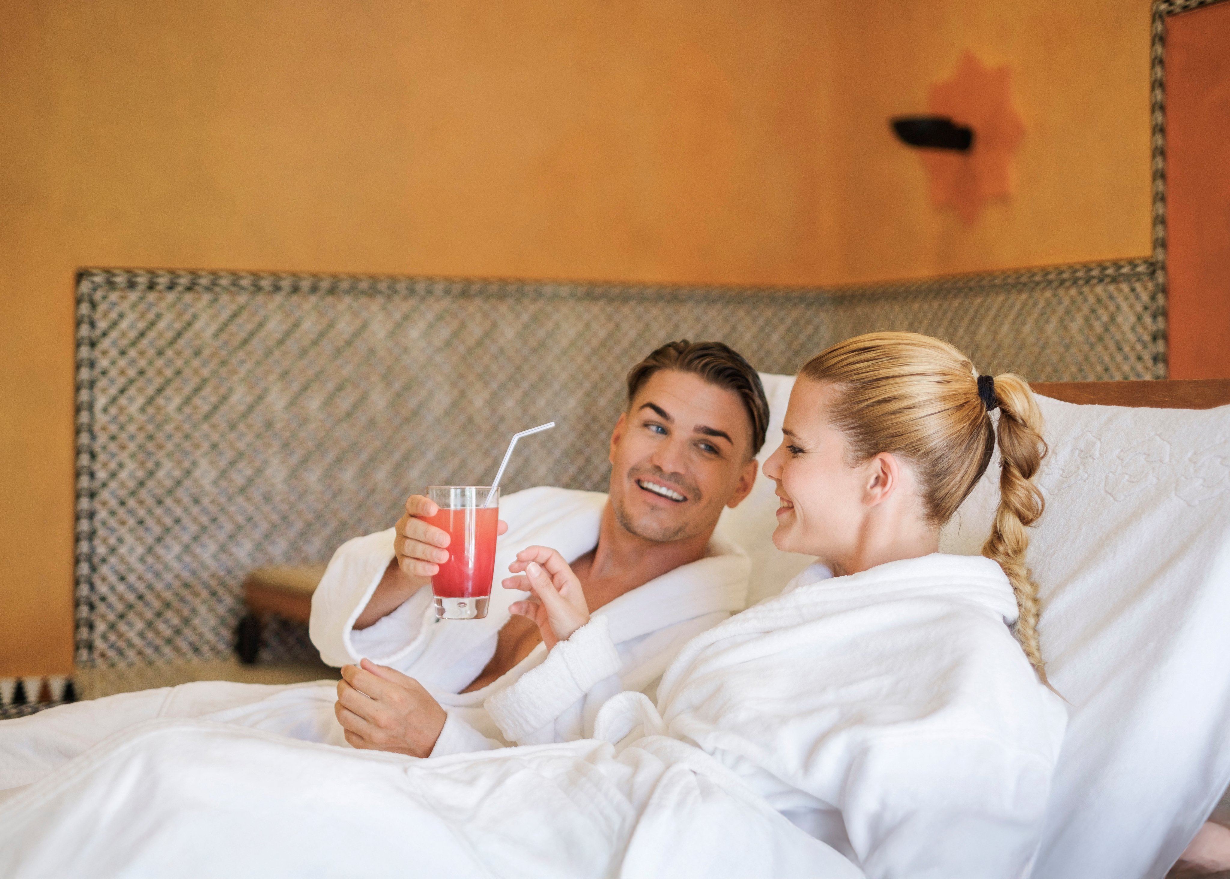 Zwei Frauen im Hotelzimmer mit Bademänteln, eine trinkt einen Cocktail, entspannte Atmosphäre