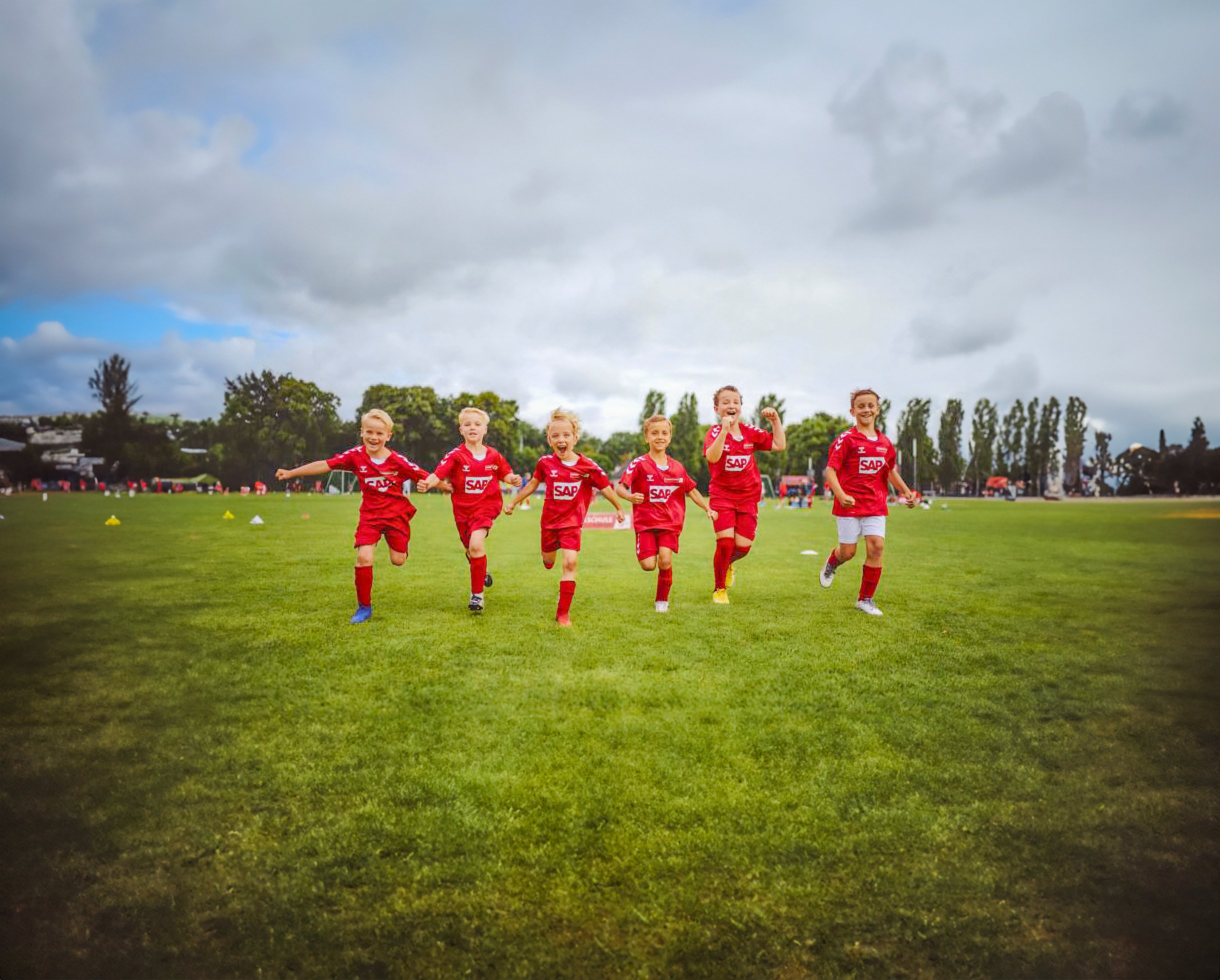 Jugendequipe trainiert auf grünem Fußballfeld, im Hintergrund Bäume und Wolkenhimmel.