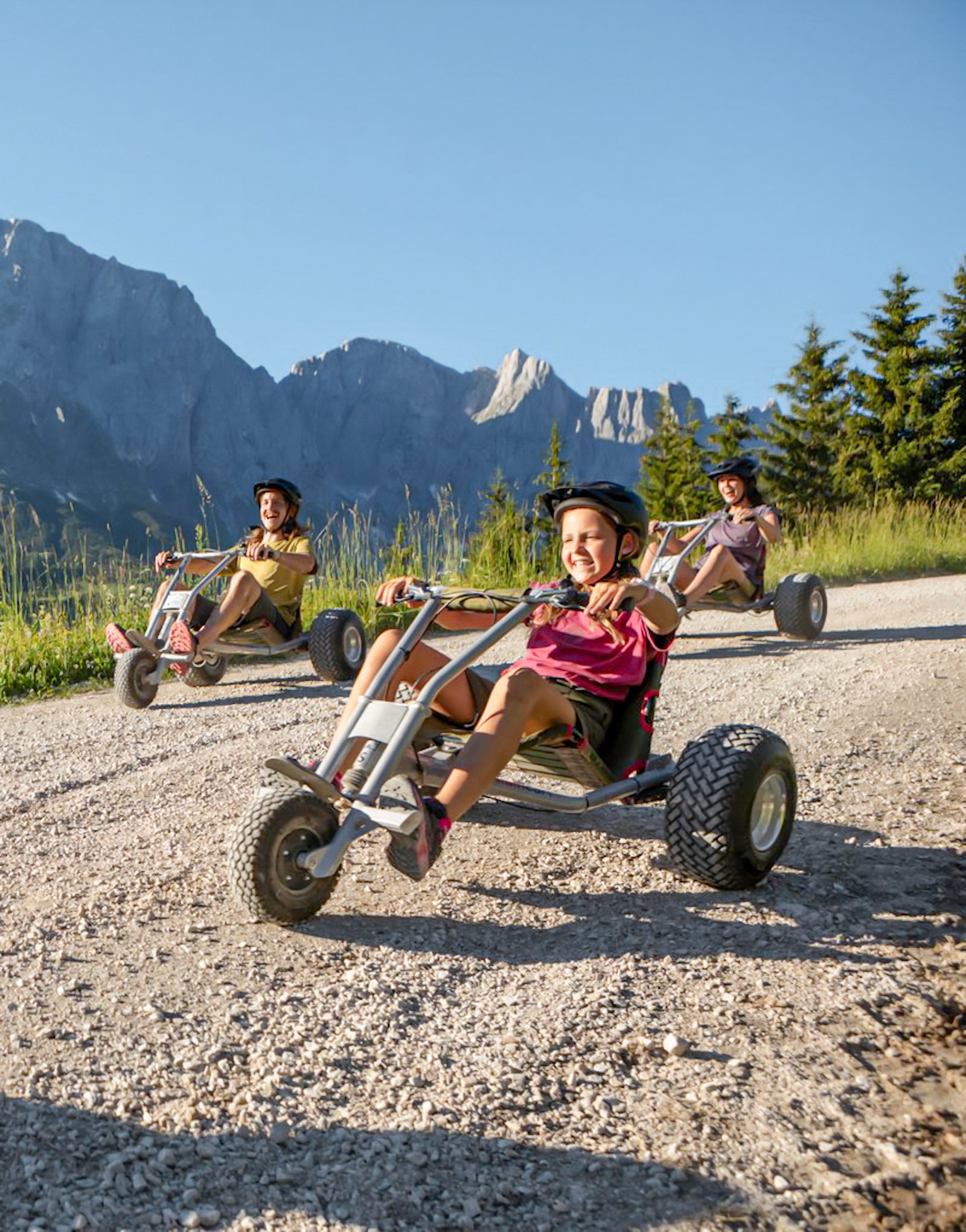 Drei Kinder fahren auf Pedalhidern in der Natur mit Bergen im Hintergrund