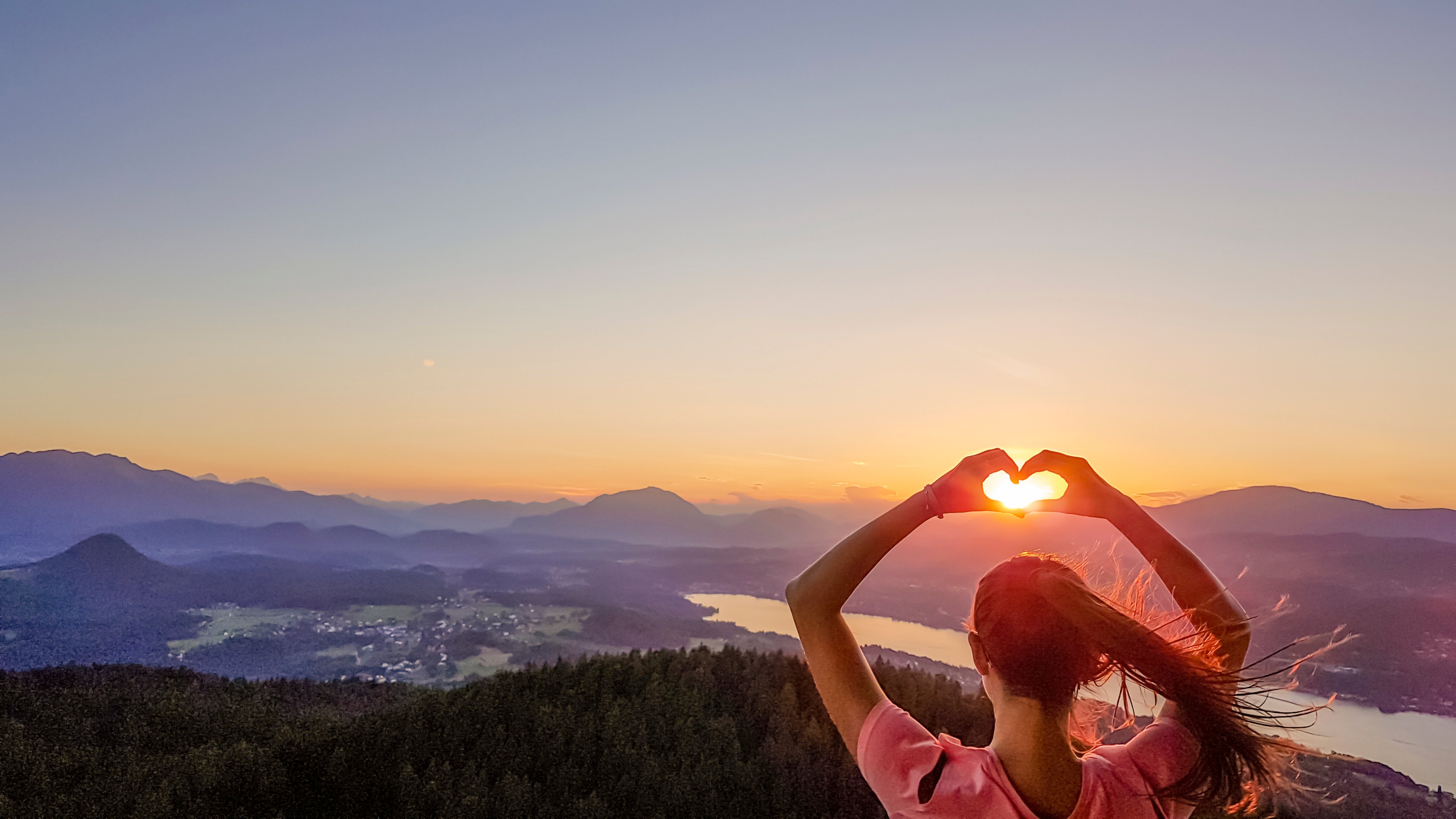 Frau bei Sonnenuntergang macht Herzzeichen mit Händen vor Seen- und Bergkulisse