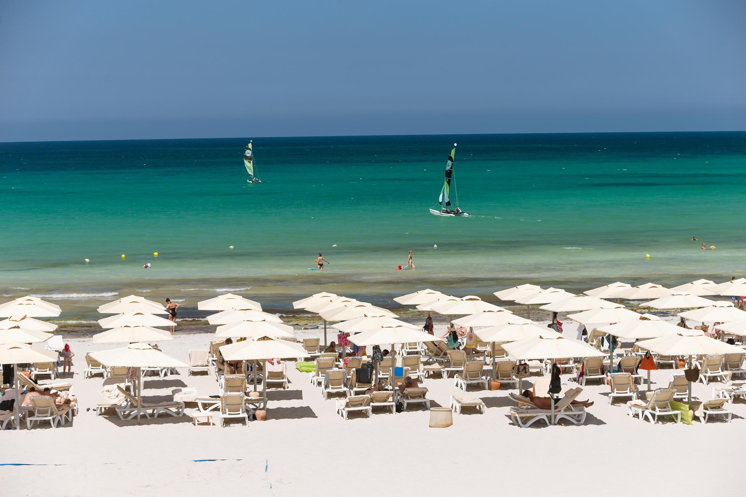 Blick auf einen Strand mit Sonnenschirmen, Liegen und Segelbooten im blauen Meer